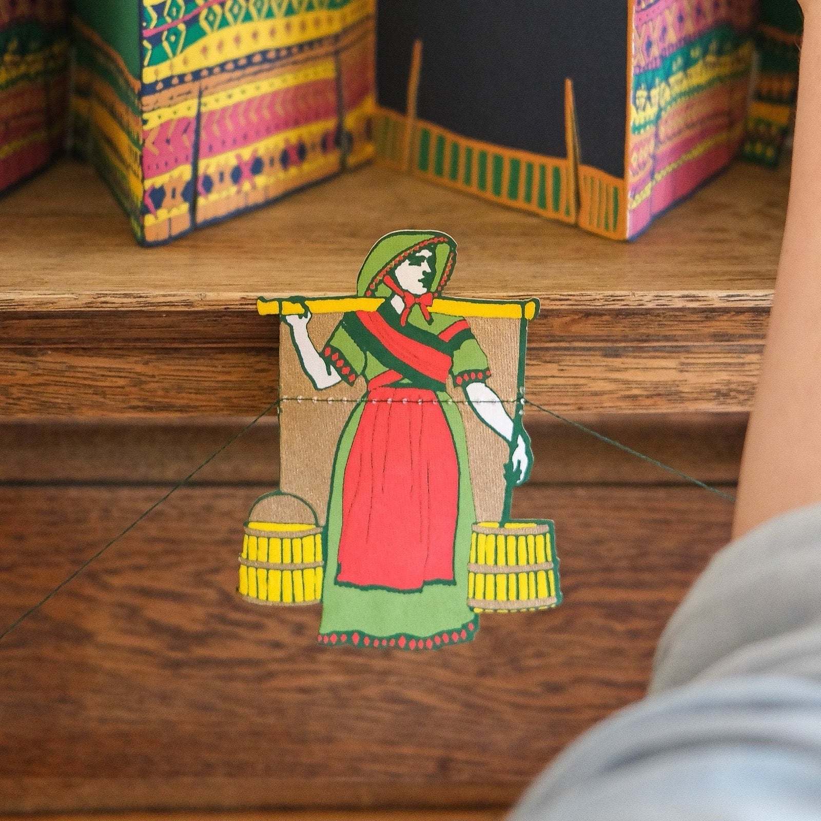 Colourful paper cutout of a woman with baskets on a wooden surface.