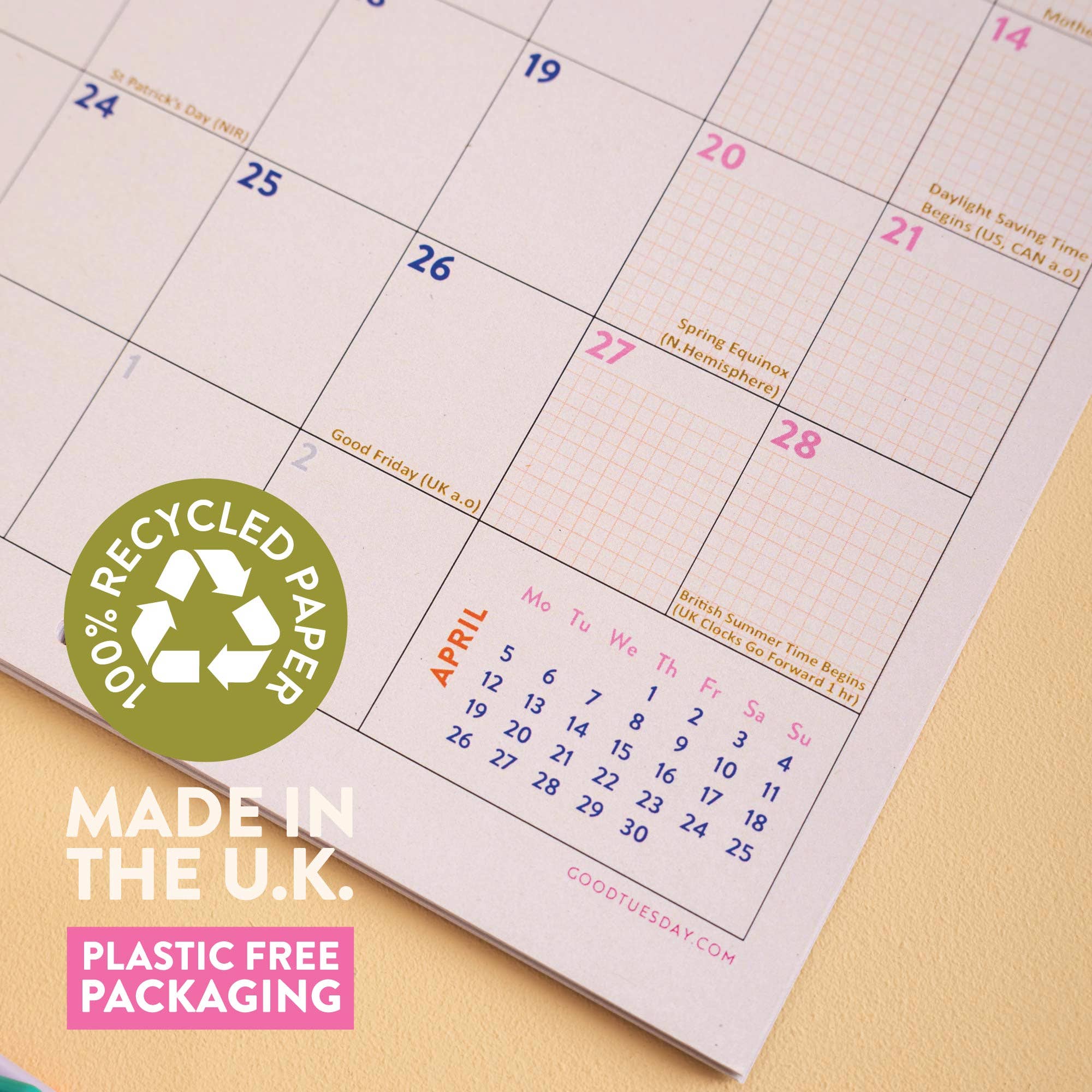A close-up of a paper calendar page showing April with pink grid squares, blue weekday numbers, and small printed notes. A green circular badge says '100% Recycled Paper,' with additional text 'MADE IN THE U.