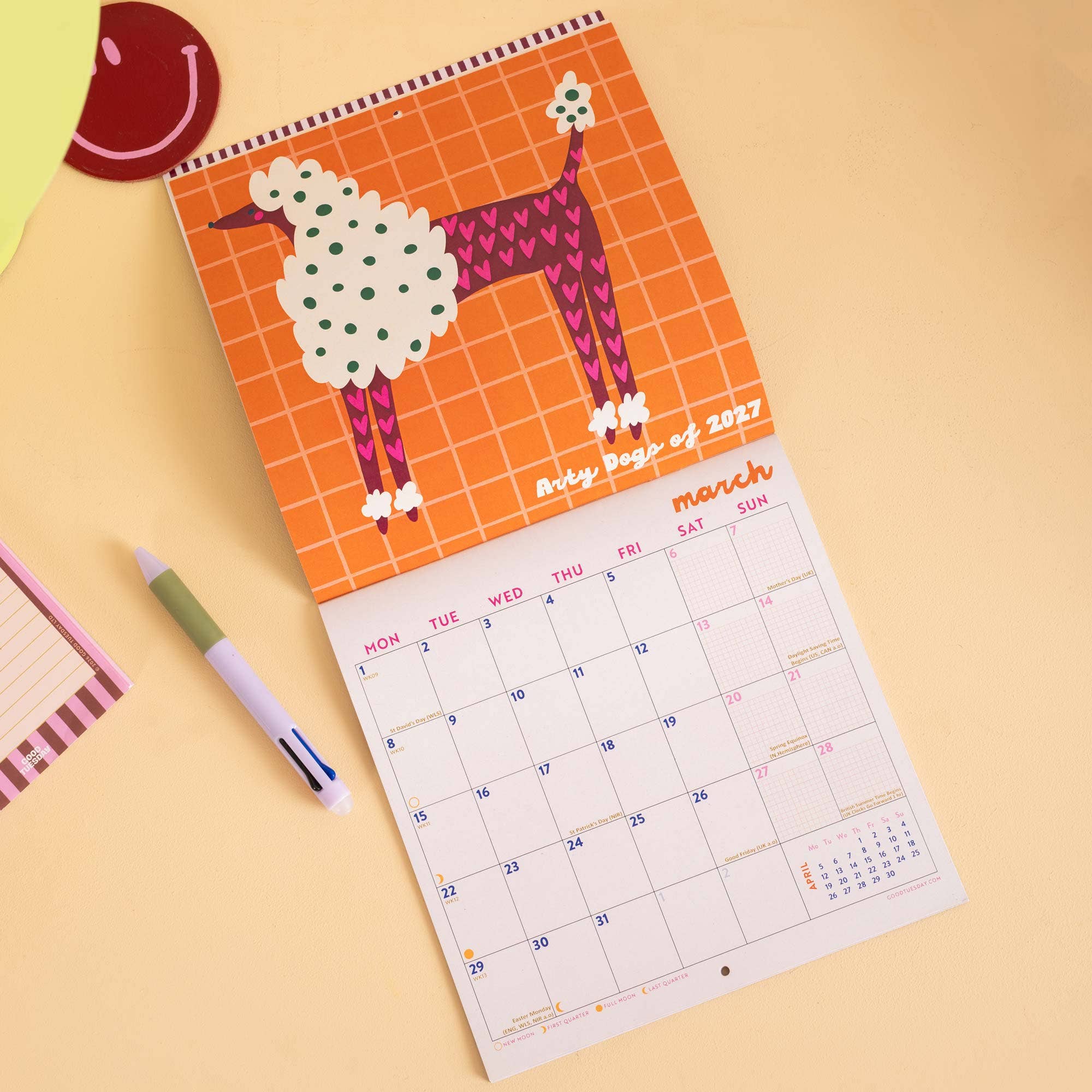 A spiral-bound monthly desk calendar for March is displayed on a warm beige surface. The cover features a stylised poodle illustration in orange, white, green, and pink.
