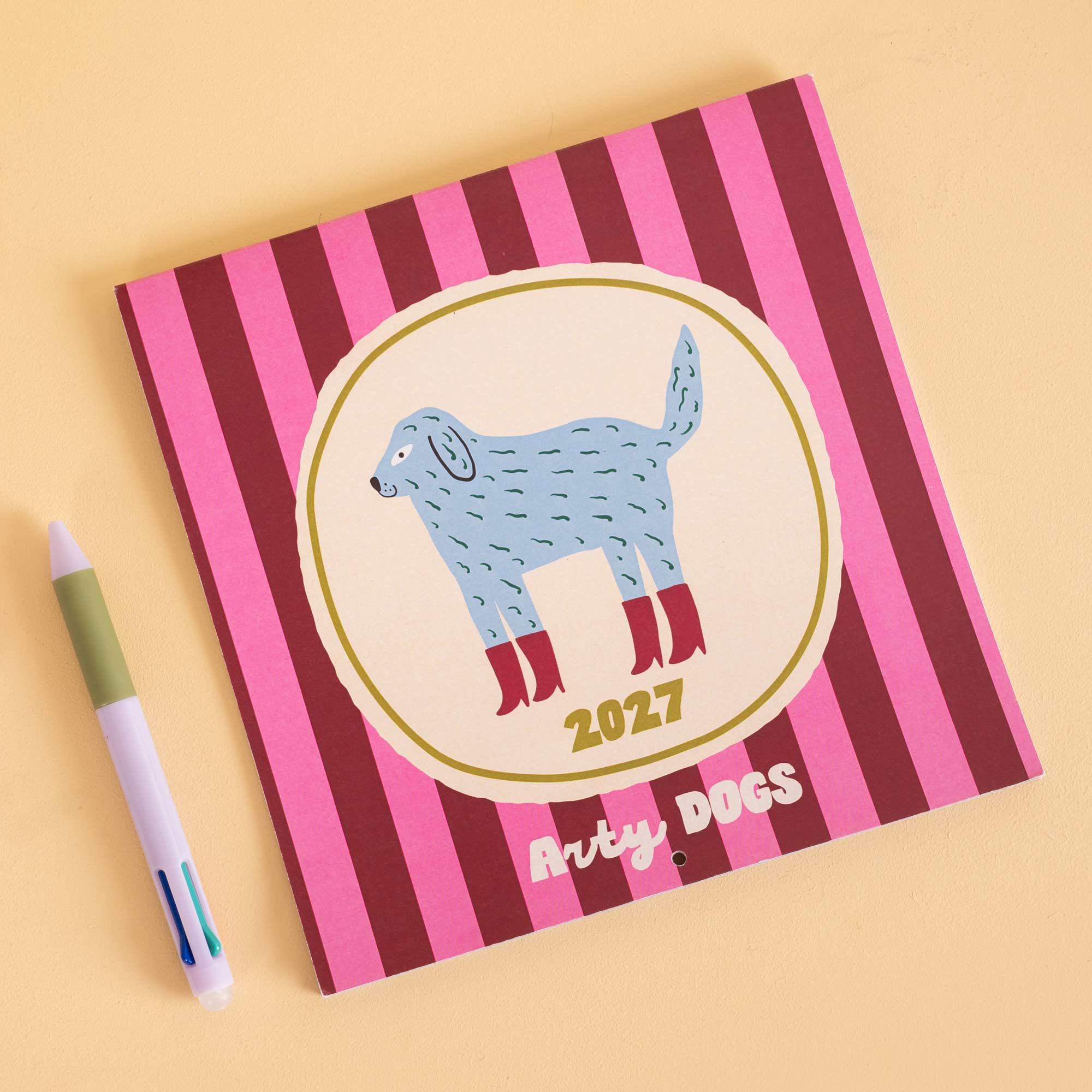 A square hardcover calendar with pink and dark red vertical stripes, featuring a central illustration of a light blue dog with red boots inside a gold oval, with the text 'Arty DOGS' and '2027' on the cover. A white and green pen rests to the left on a warm beige background.