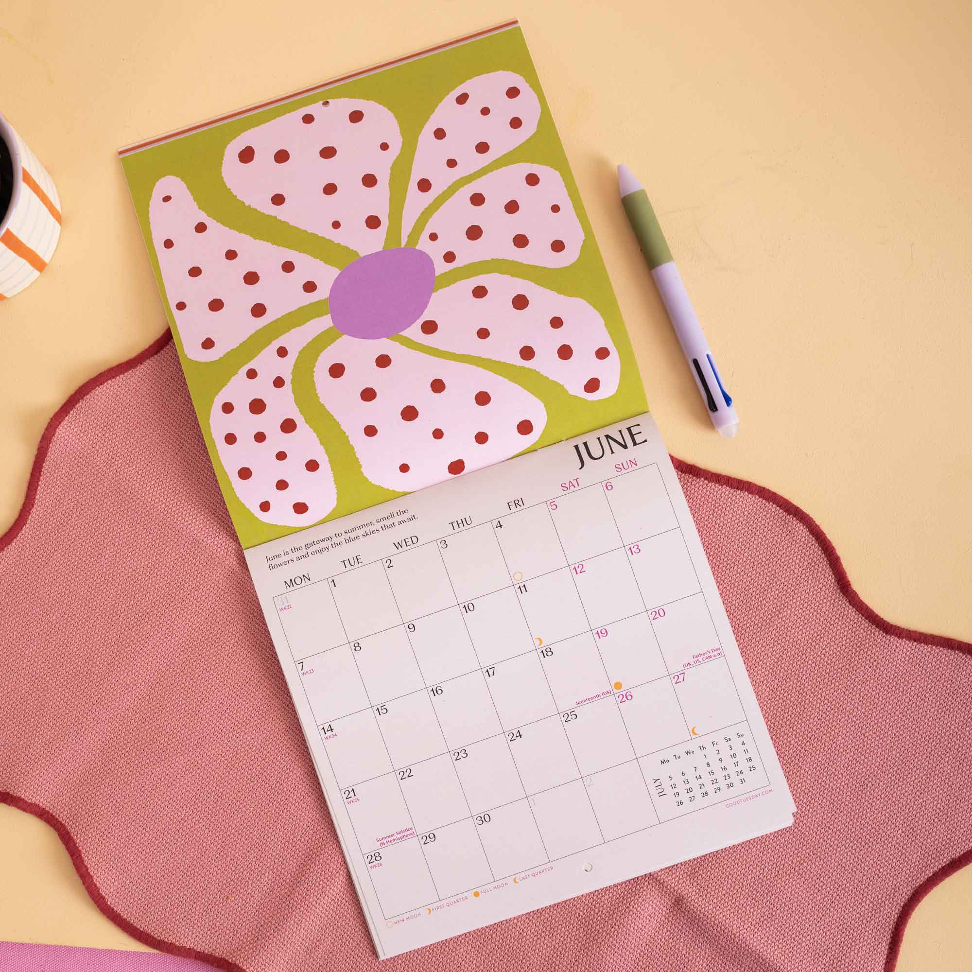 A spring-themed desk calendar featuring a large pink flower with red polka dots on an olive green background, with the June calendar page visible underneath. The calendar is styled on a pink scalloped fabric surface with a green pen nearby on a warm beige background.