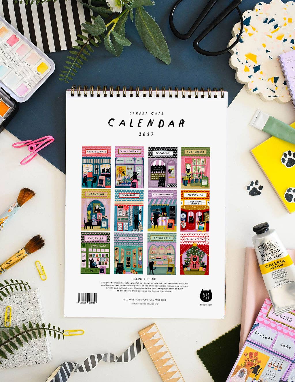 A spiral-bound 2023 calendar titled 'Street Cats Calendar' on a white and blue surface. The calendar cover features a grid of colourful illustrations depicting various cat-themed storefronts.