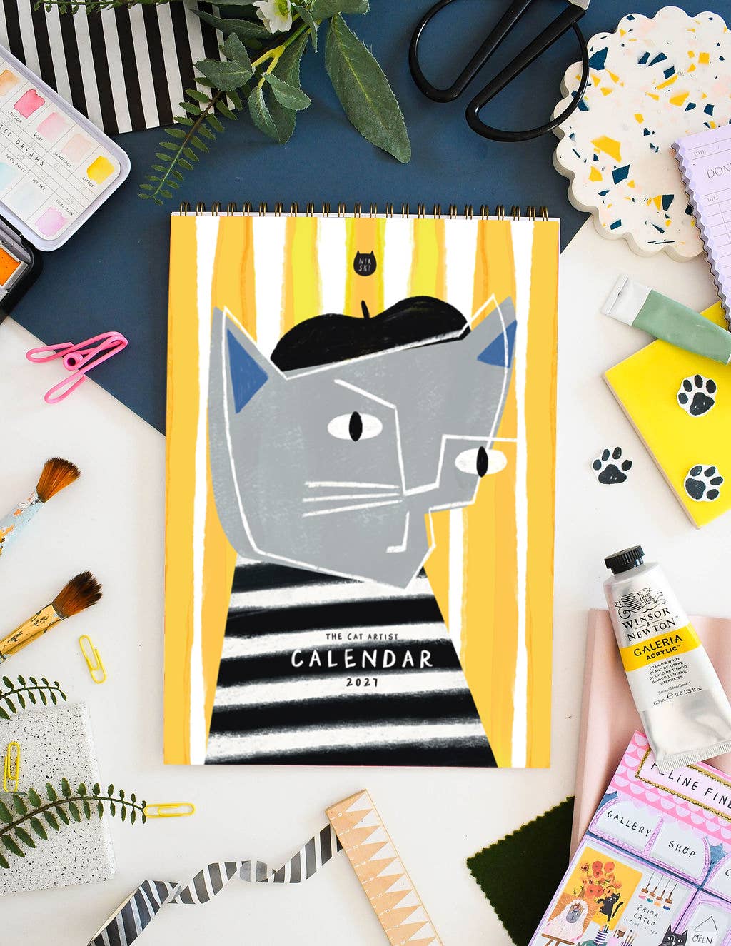 A spiral-bound 2021 art calendar featuring a grey cat in a black beret and striped shirt on a yellow-and-white striped background, styled in a flat lay with art supplies around it.