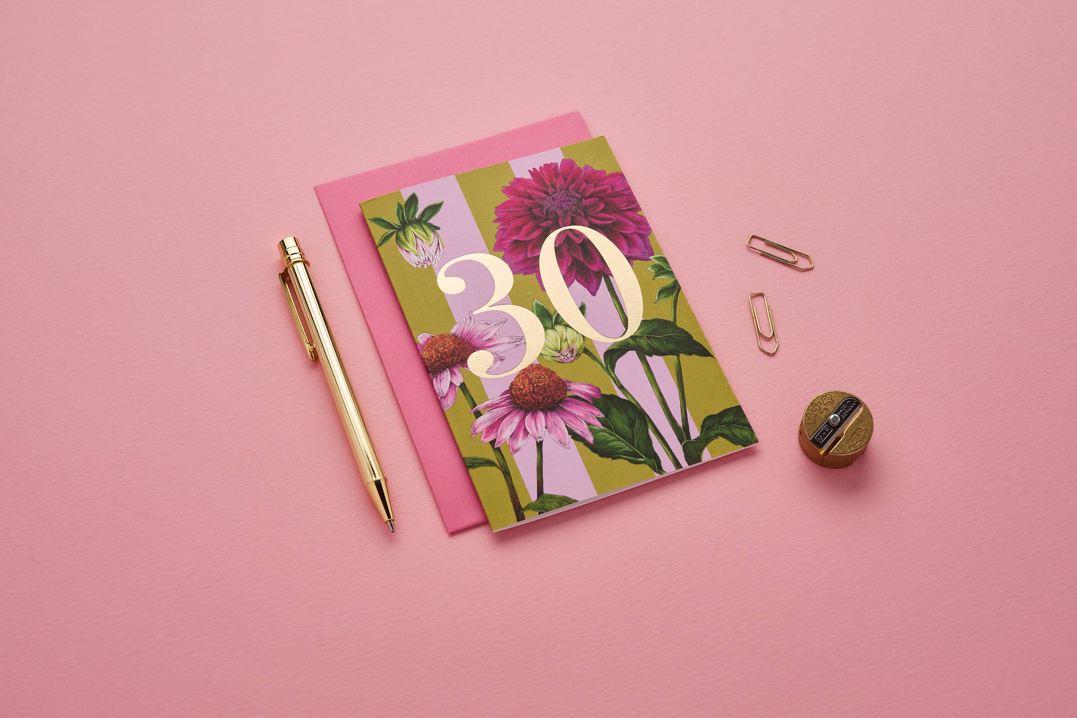 Notebook with floral design and '30' on a pink background, accompanied by a pen and paper clips.