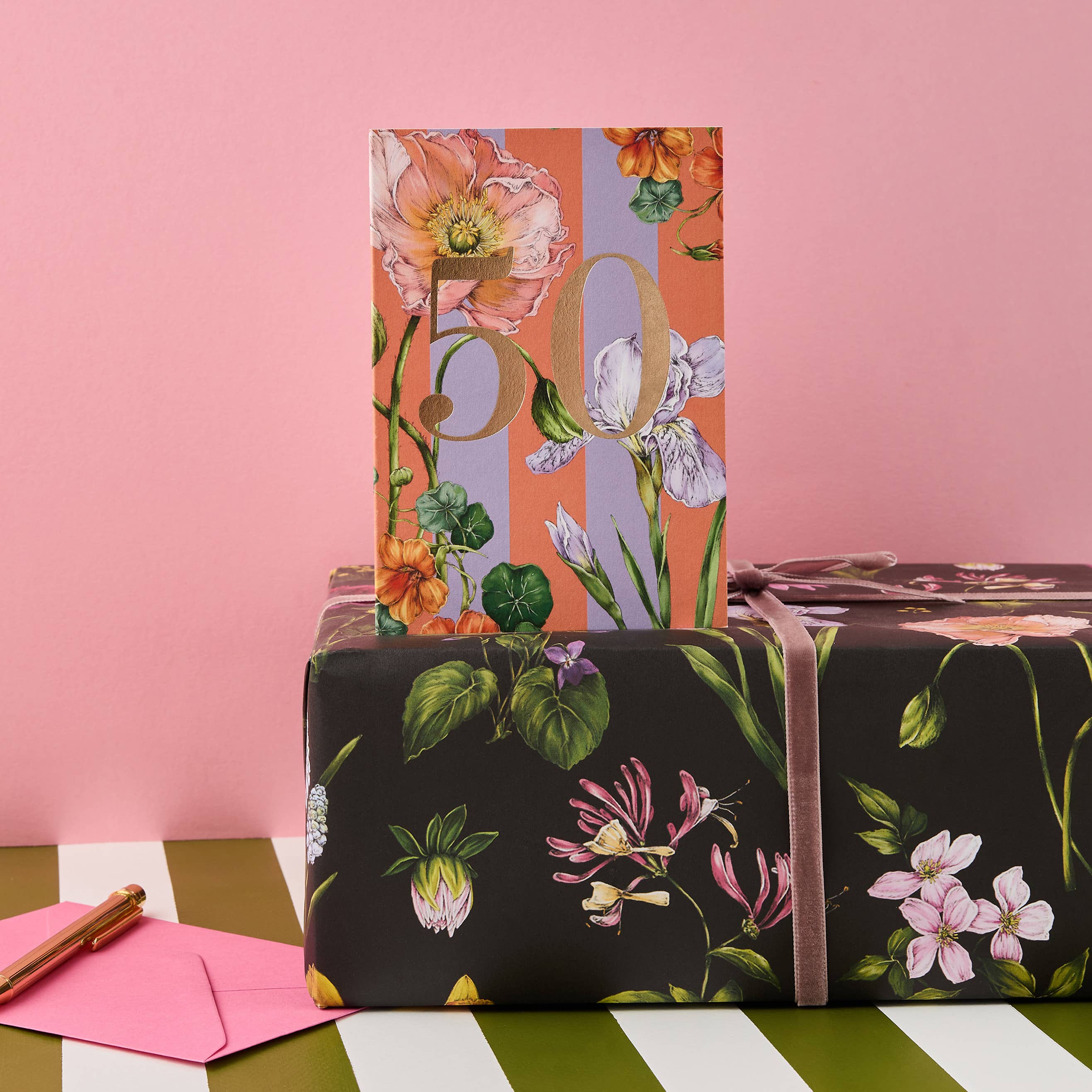 Gift wrapped in floral paper with a pink envelope on a striped surface against a pink background.