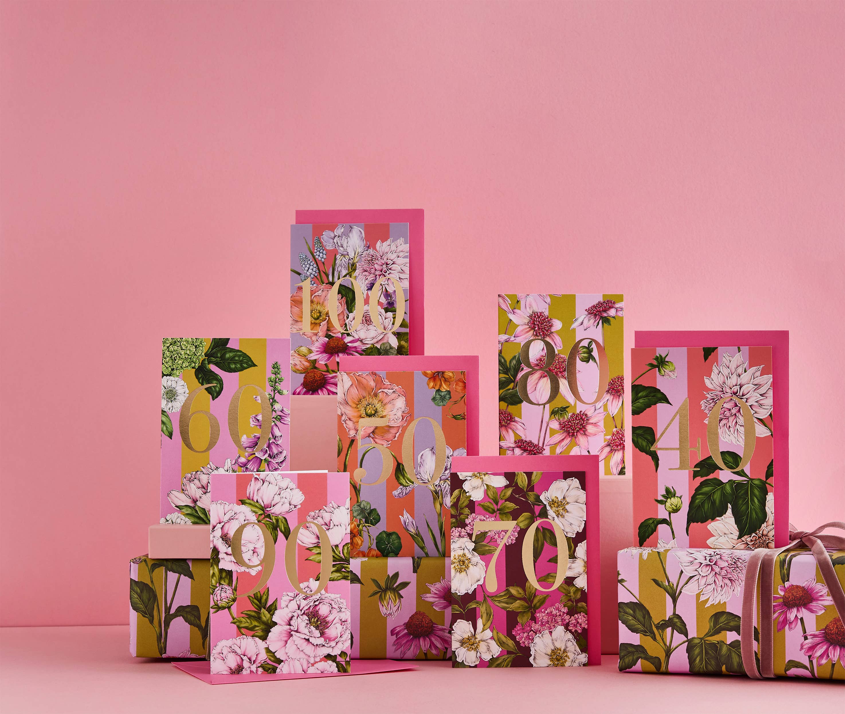 A collection of decorative gift boxes and cards with a vibrant floral and botanical print in pink, green, yellow, and white, arranged on a matching pink surface and background.
