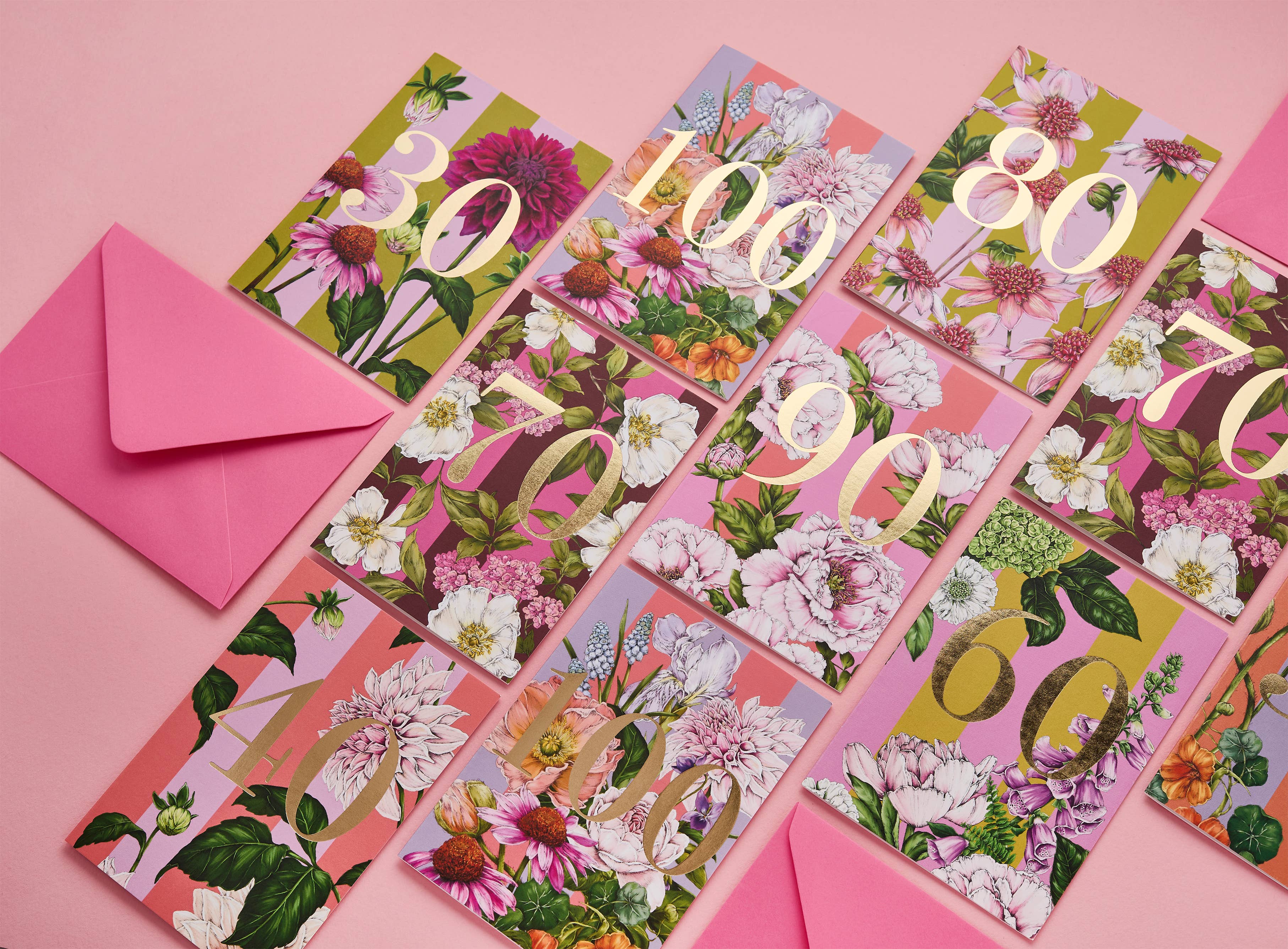 A set of floral milestone cards arranged on a pink background, each card featuring a different gold number from 30 to 80+ printed over a pink, green, and white botanical pattern. A matching pink envelope is placed to the left.