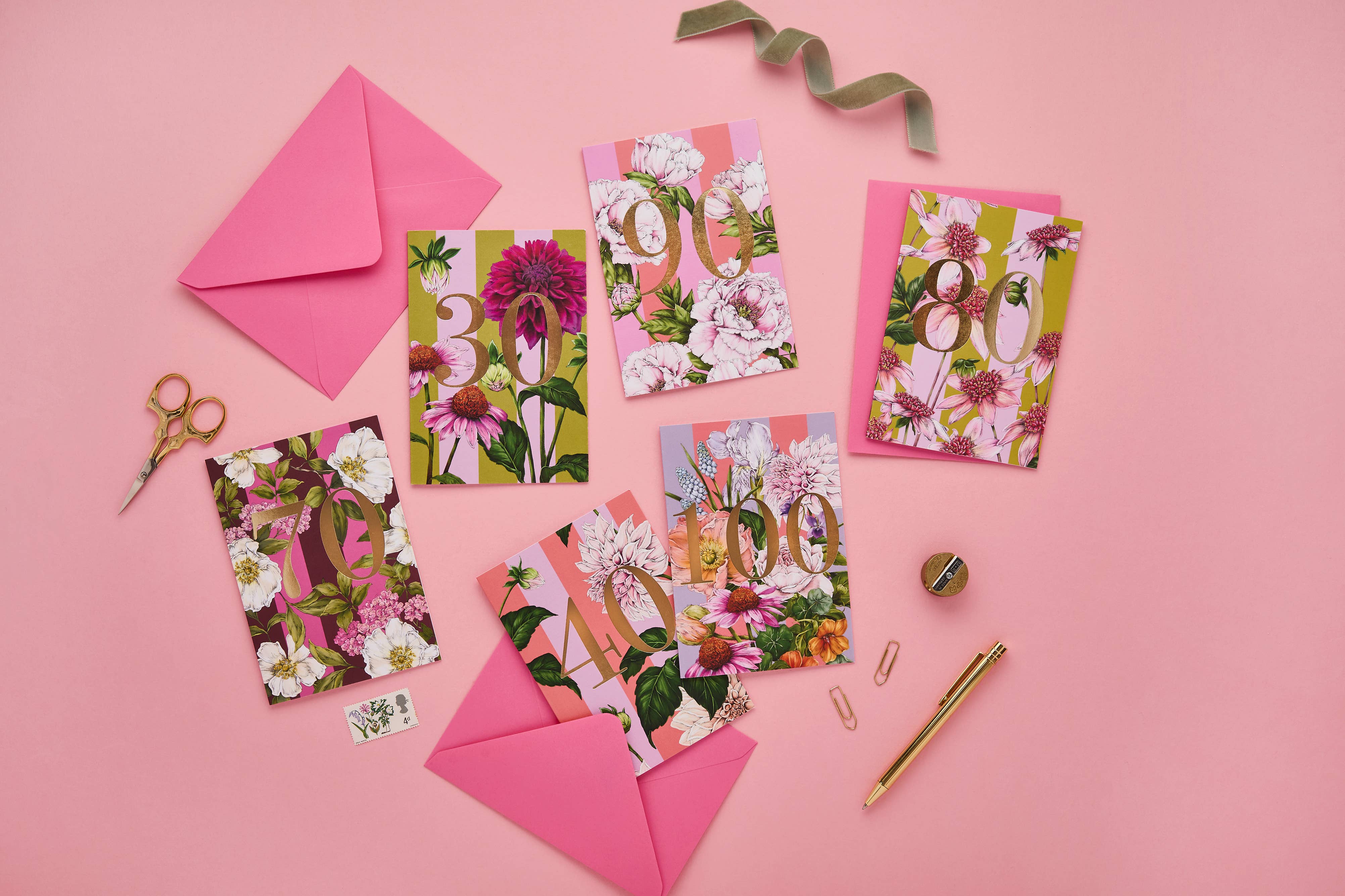 A styled flat-lay of floral greeting cards with matching pink envelopes, scissors, paper clips, a pen, and ribbon on a solid pink background.