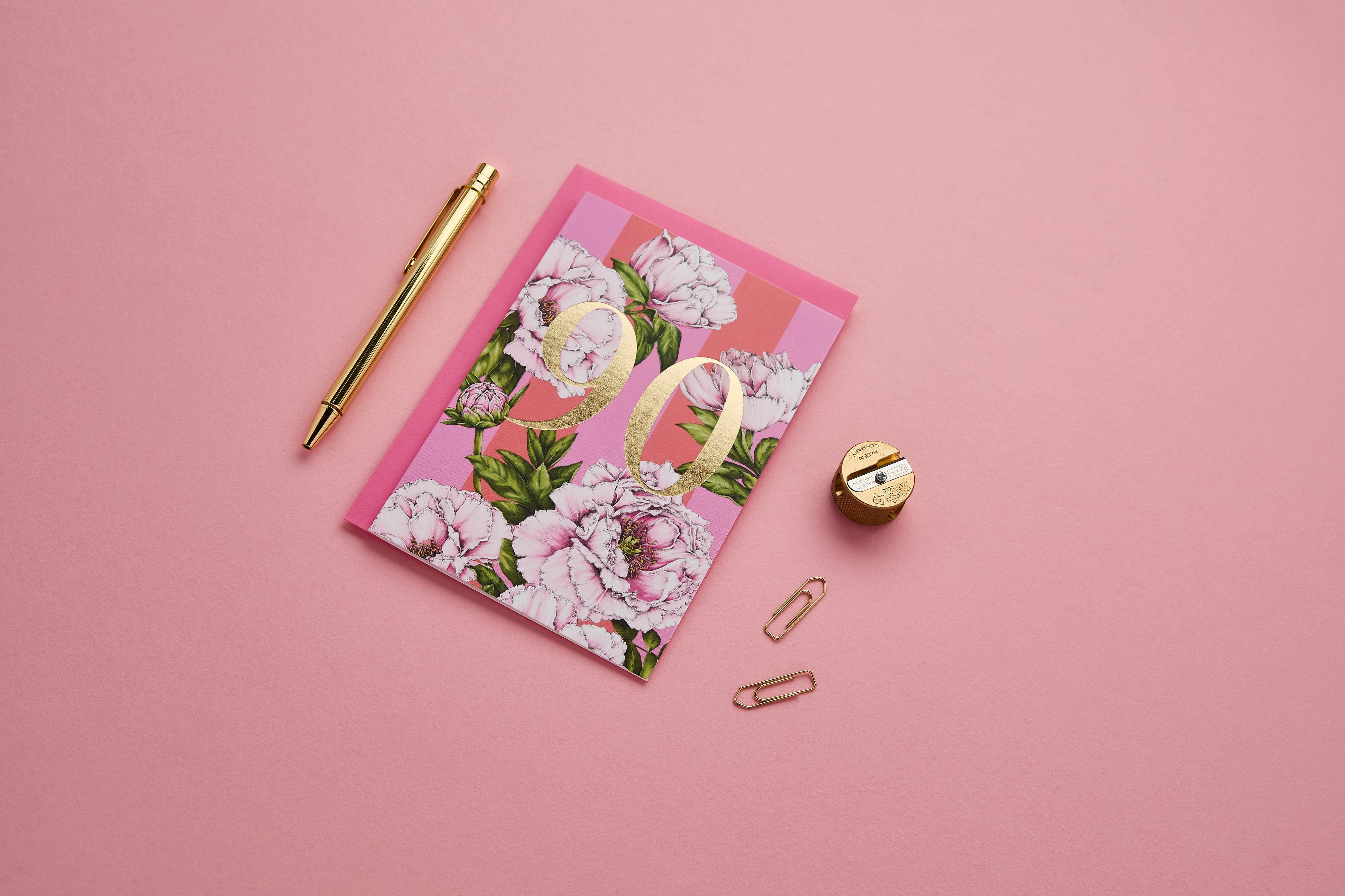 A pink floral notebook with a large gold number 90 on the cover, styled on a pink background with a gold pen, paper clips, and a small gold pencil sharpener.