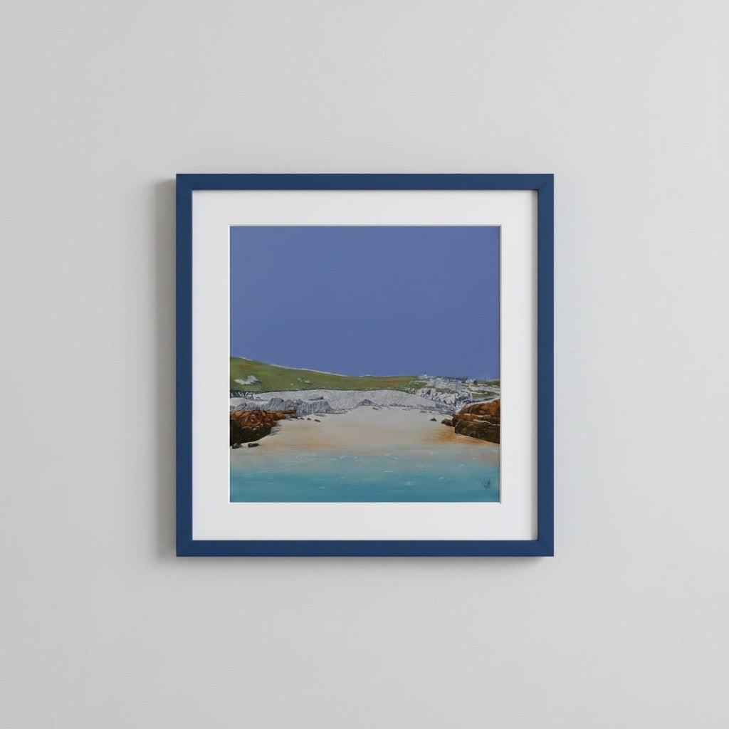 A framed coastal landscape art print featuring a sandy beach, turquoise water, green hills, and a blue sky, presented in a navy blue frame with a white mounting against a light grey wall.