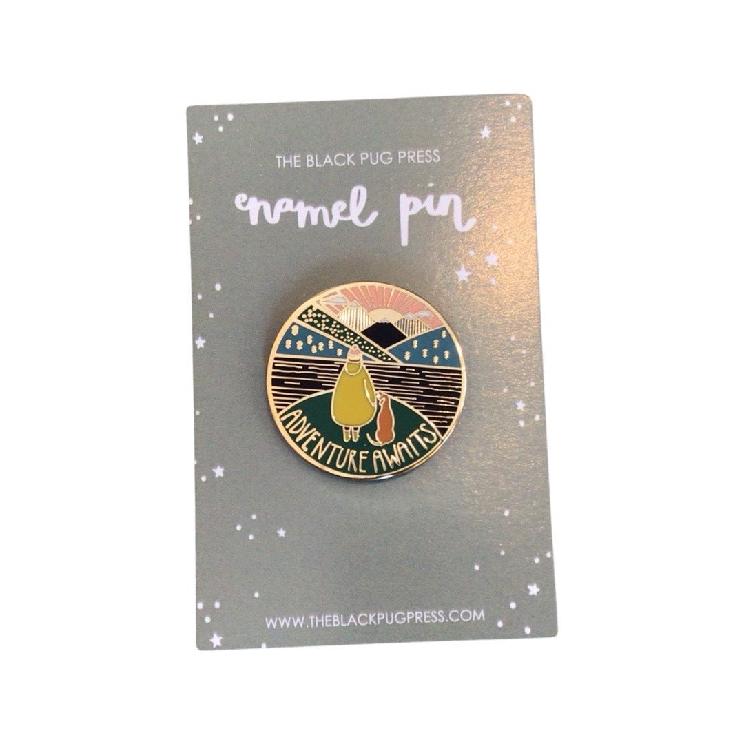 Enamel pin badge featuring an illustration of a dog and owner with a scenic background of a lake and mountains, with the text 'Adventure Awaits' on backing card.