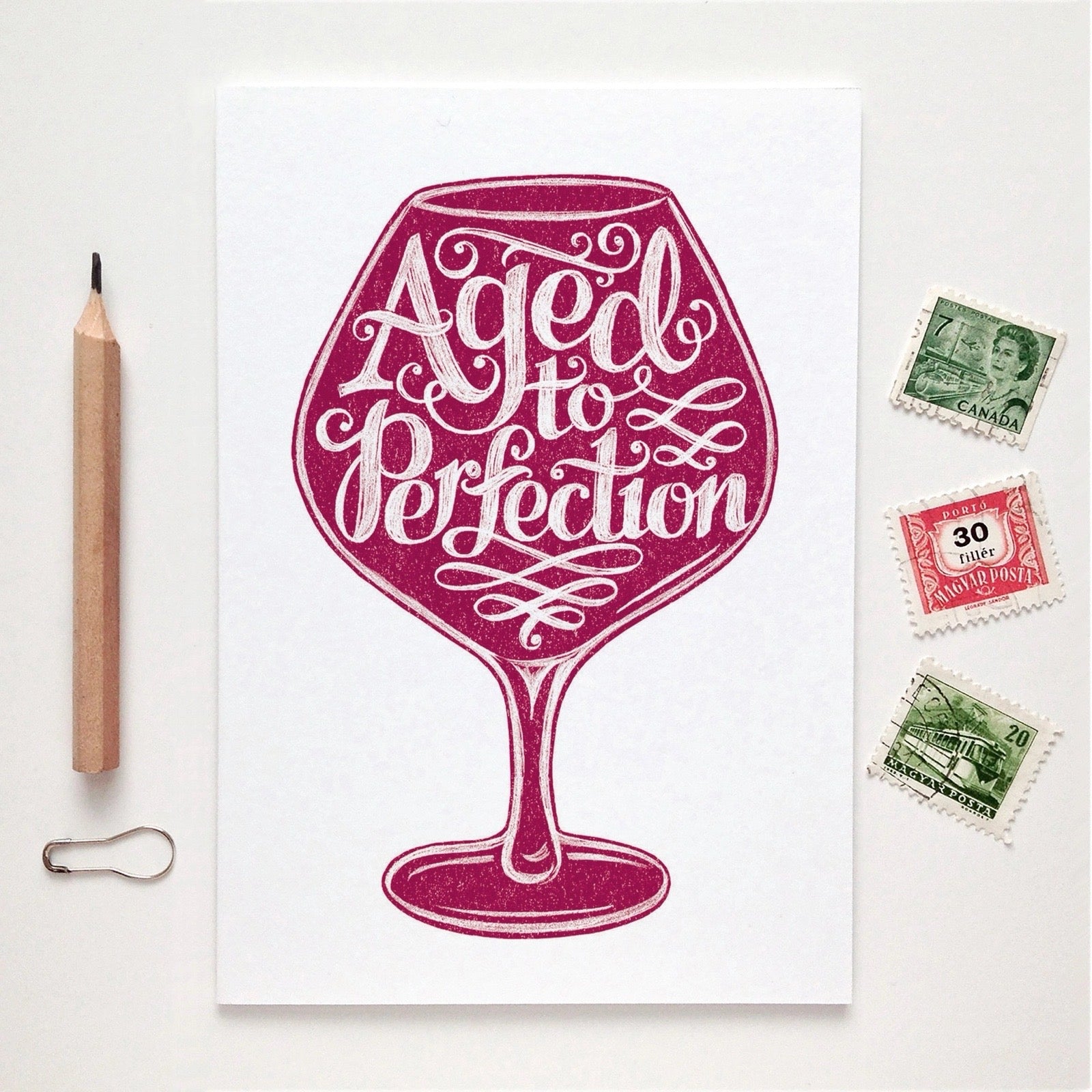 Card with a wine glass design and 'Aged to Perfection' text on a white background with stamps and a pencil.
