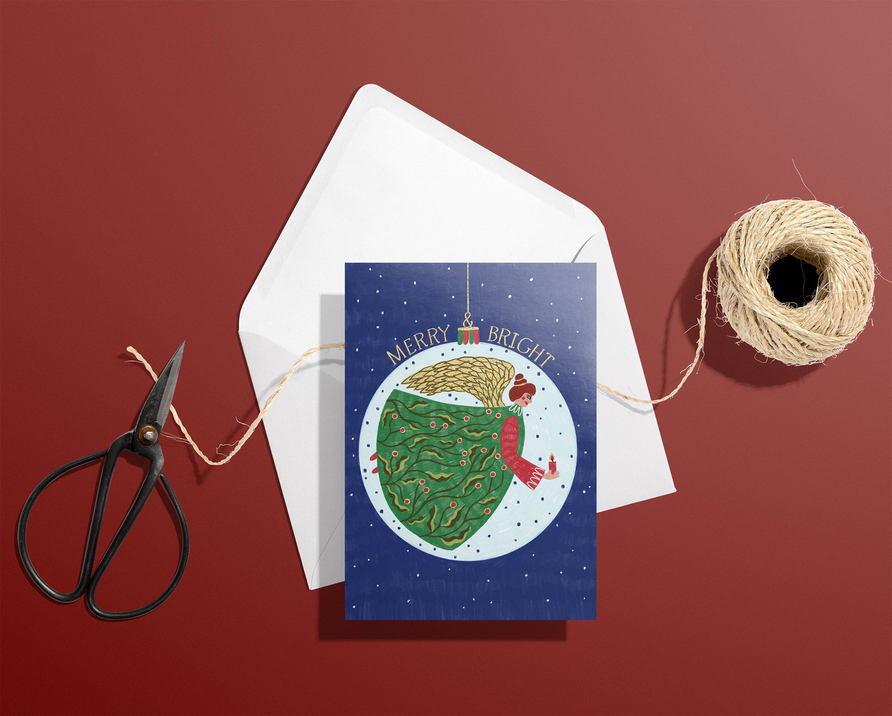A greeting card with a festive illustration of a woman in a green dress and red scarf under a gold wreath, set against a dark blue starry background with the text 'MERRY & BIGHT'. The card is displayed with a white envelope, black scissors, and a spool of twine on a solid dark red background.