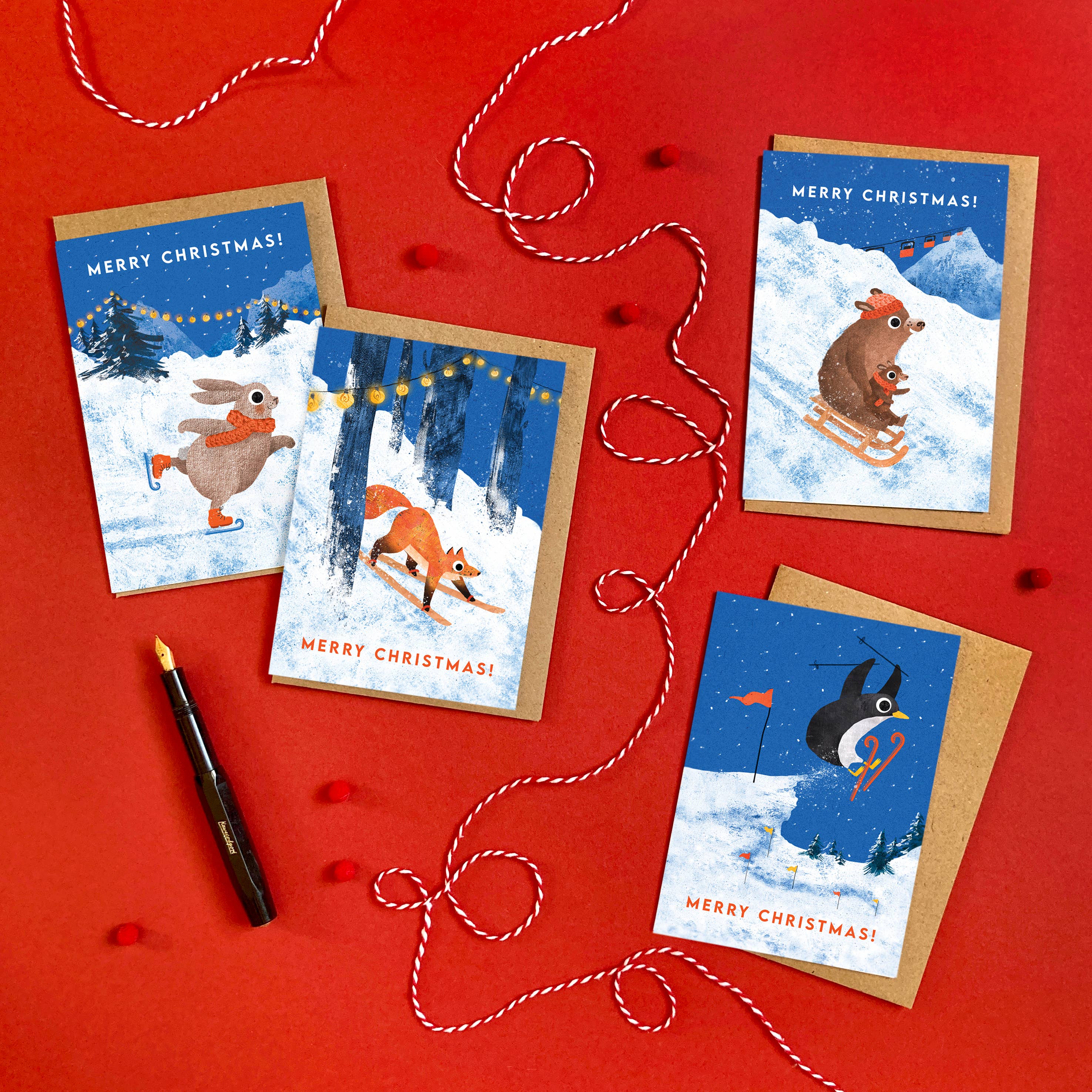 Four Christmas greeting cards with winter animal illustrations are arranged on a red background with red-and-white string, a black pen, and kraft envelopes. The cards read 'MERRY CHRISTMAS!' and show a rabbit, fox, mole, and penguin in snowy scenes.