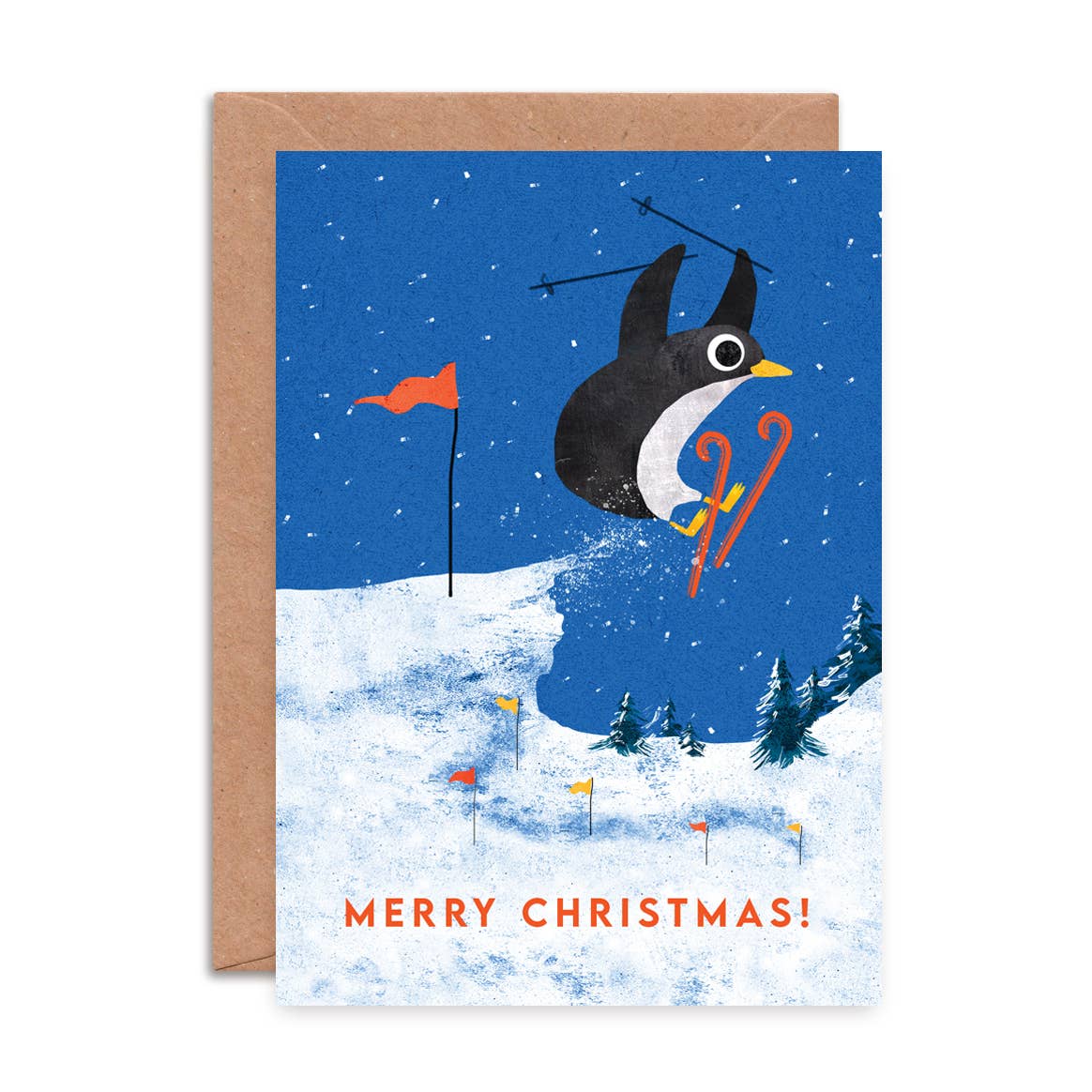 A Christmas greeting card featuring a penguin skiing through the snow under a blue night sky, with the text 'MERRY CHRISTMAS!' at the bottom, shown with a brown kraft envelope behind it on a white background.