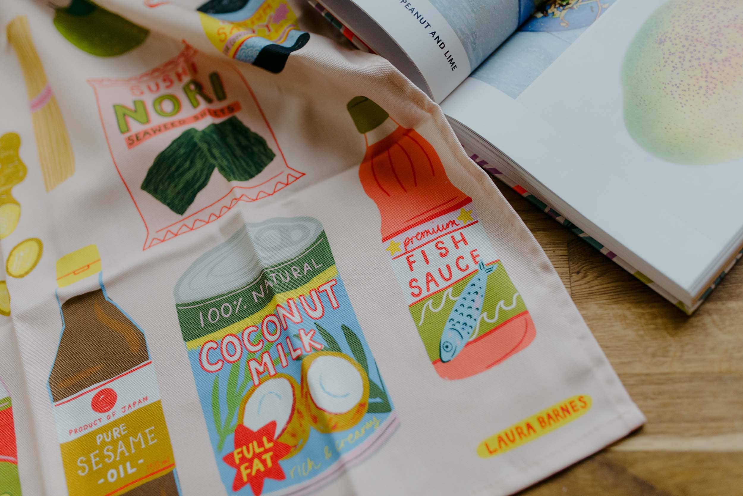 Tea towel with illustrated food products and branding on a wooden surface.