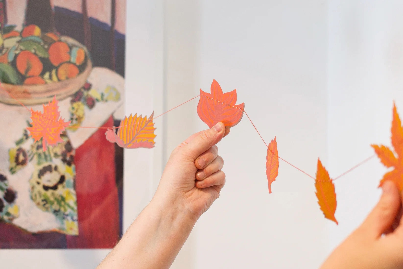 Person holding a string of orange paper leaves in front of a painting.