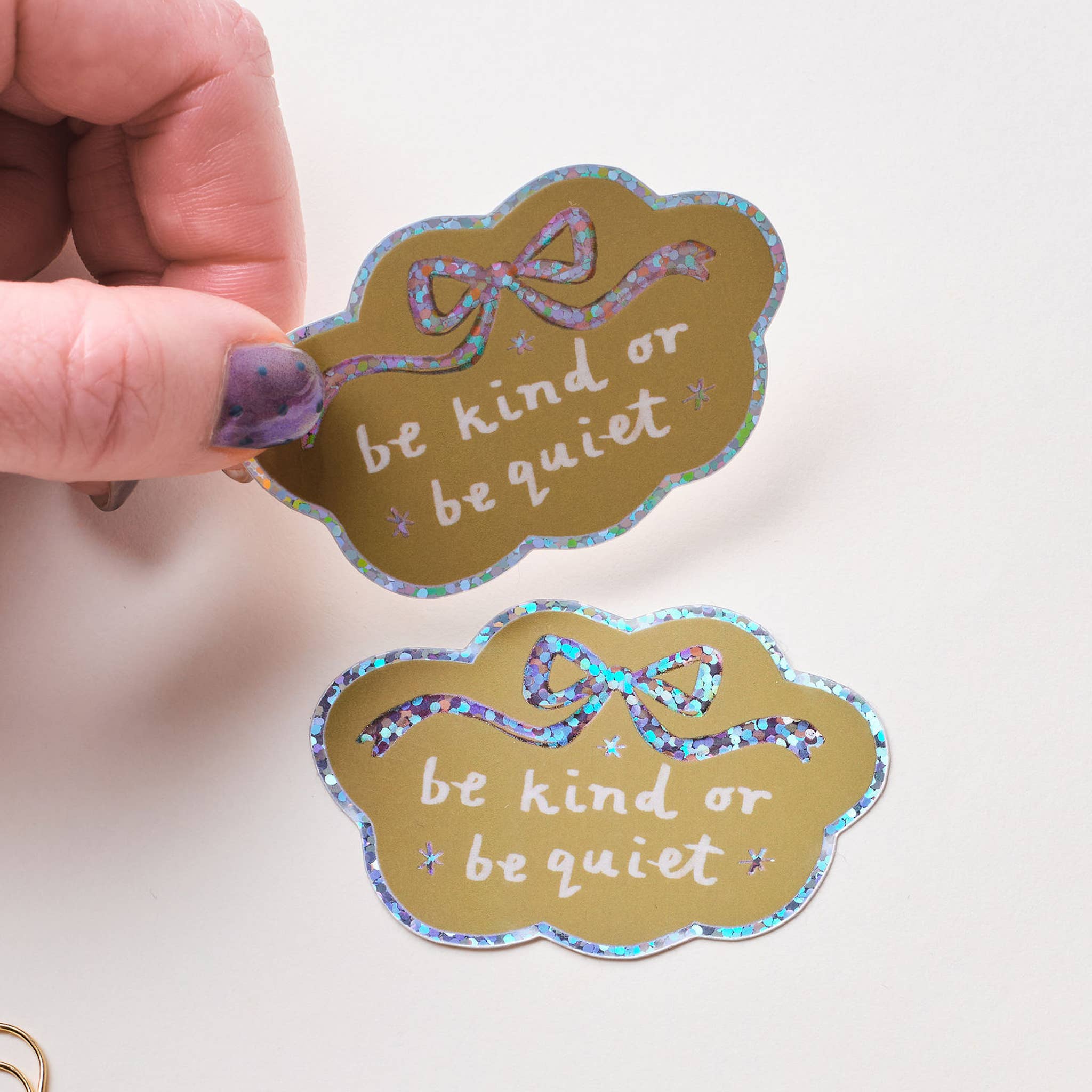 Two gold stickers with 'be kind or be quiet' text held by a hand on a white background.