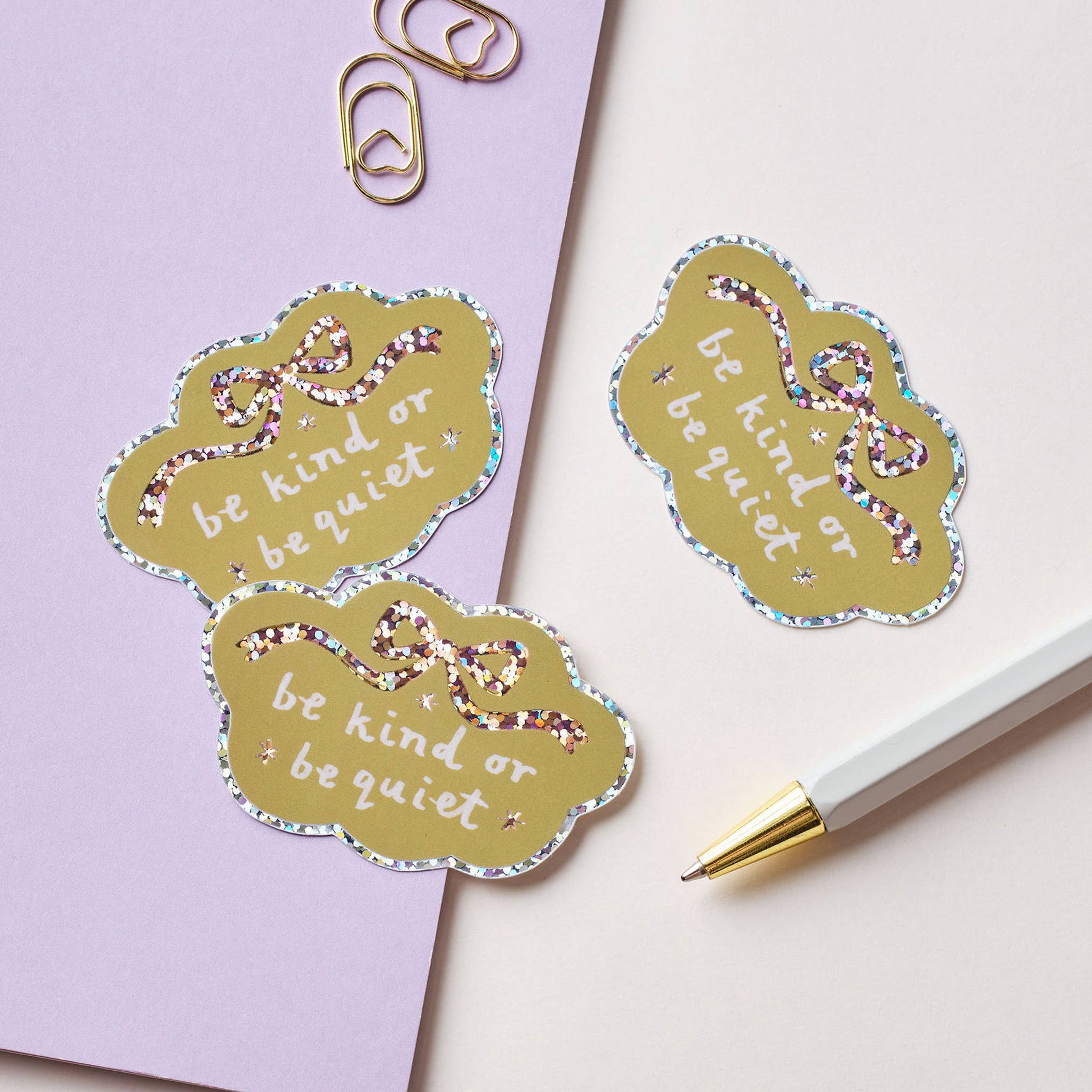 Three decorative stickers with 'be kind or be quiet' text on a light purple background with a pen.