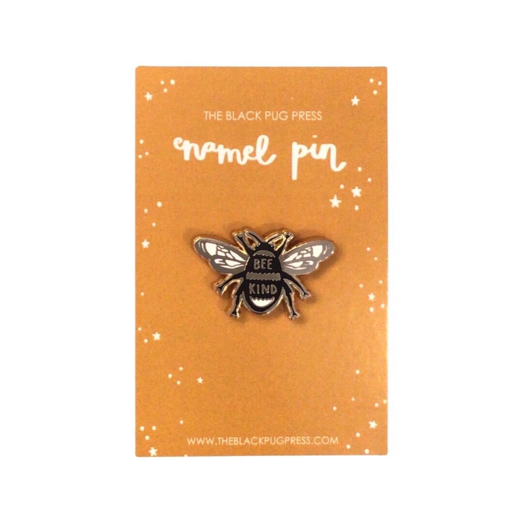 Bee-shaped enamel pin badge. in black, white and gold with the text 'Bee Kind' on backing card.