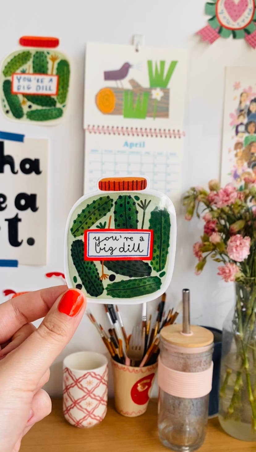 Hand holding a sticker with a container with pickles and 'you're a big dill' text, with a calendar and flowers in the background.
