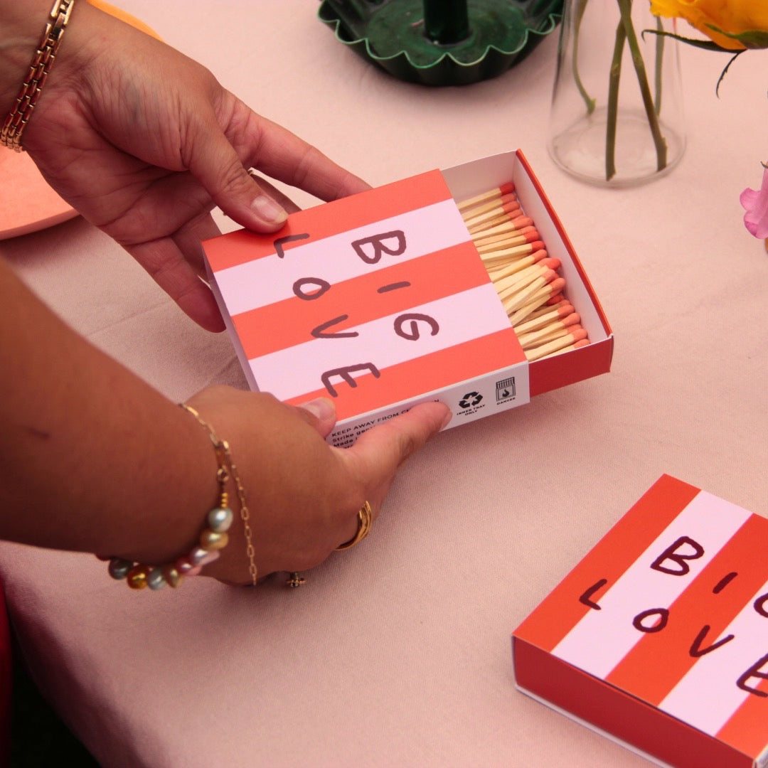 This matchbox features a red and pink striped design with the text 'Big Love', on a table.
