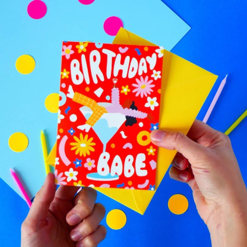 Hand holding a colorful birthday card with 'Birthday Babe' text against a blue background with polka dots.
