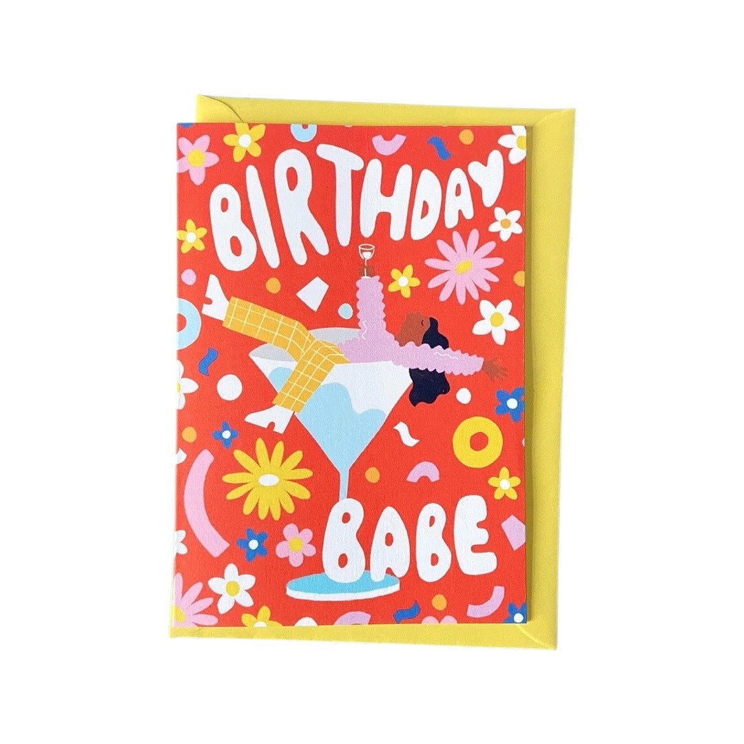 Birthday card featuring an illustration of a partied-out girl lying on top of a giant cocktail glass and the text 'Birthday Babe' in white, all on a red background with colourful flowers and shapes.
