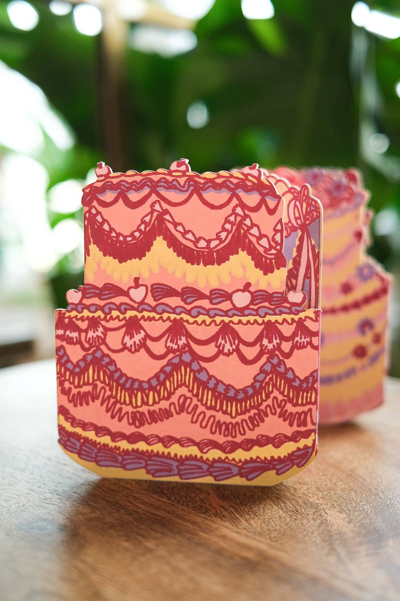 Decorative pink and yellow cake-shaped card on a wooden surface with a blurred green background.