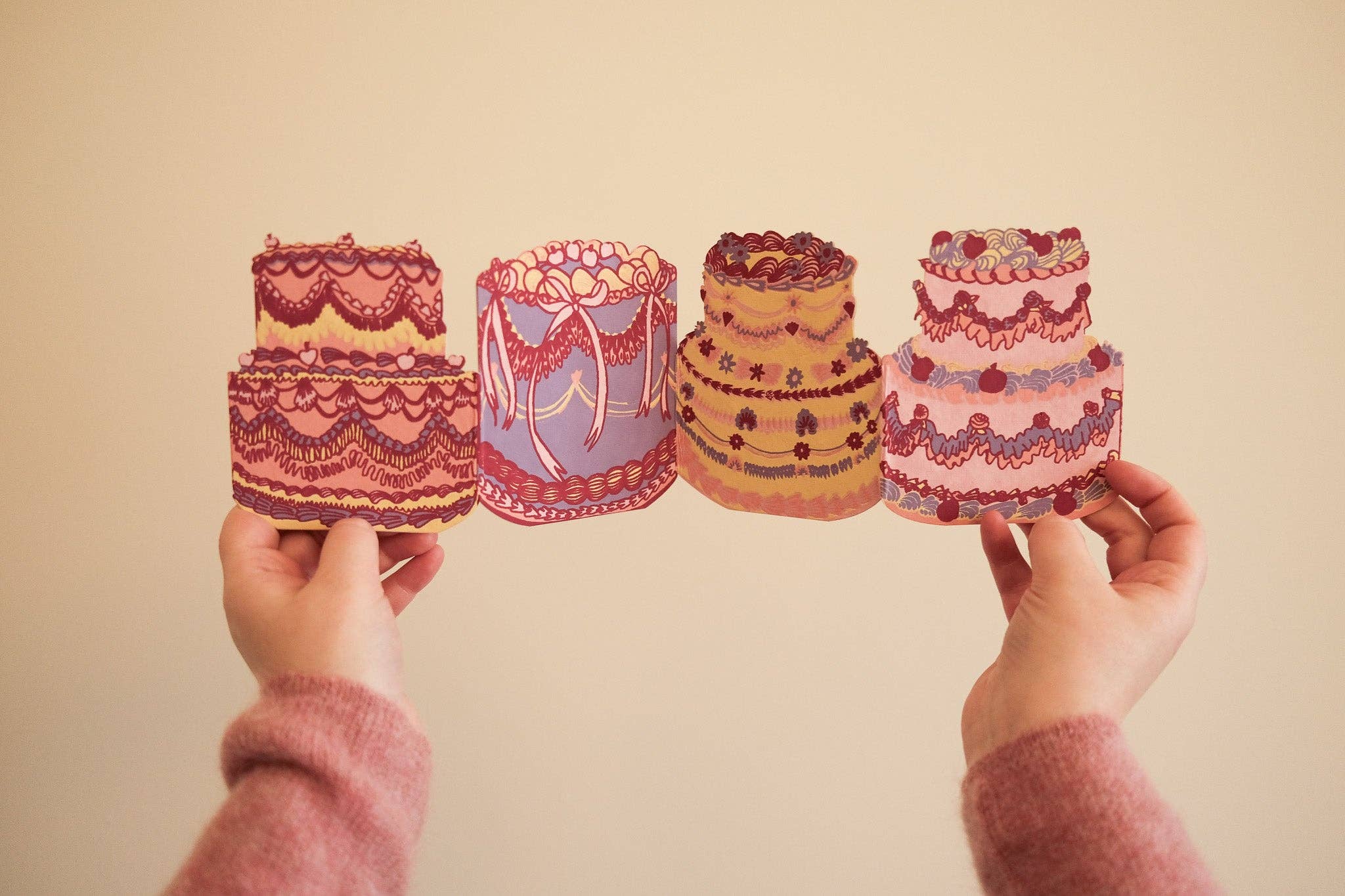 A card featuring four intricately designed cakes held by hands against a plain background.