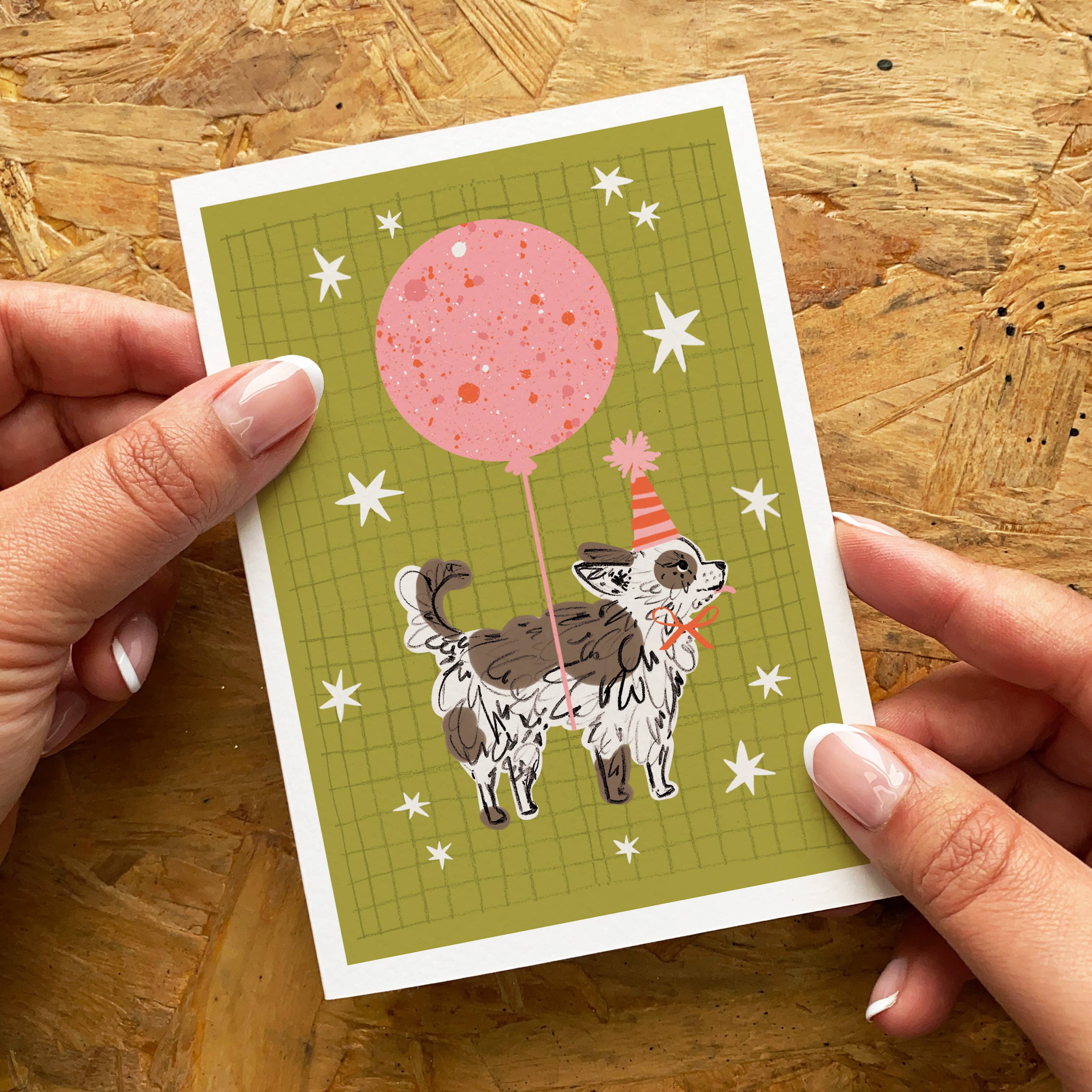 A hand-held greeting card featuring an illustration of a small dog holding a pink balloon, set against a wood-grain background. Two hands with manicured nails hold the card from the sides.