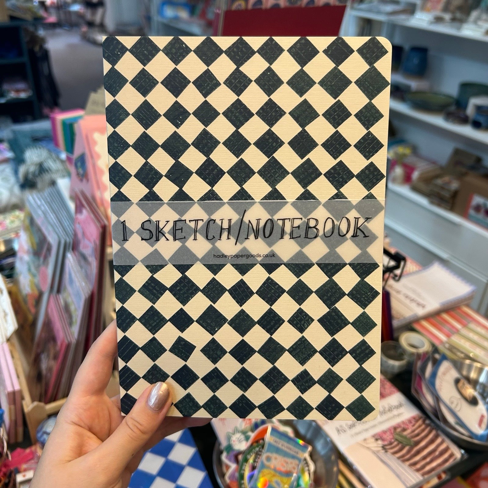 A hand holds a sketchbook or notebook with a black and beige diamond patterned cover in a store setting. A semi-transparent banner across the middle reads '1 SKETCH/NOTEBOOK'.