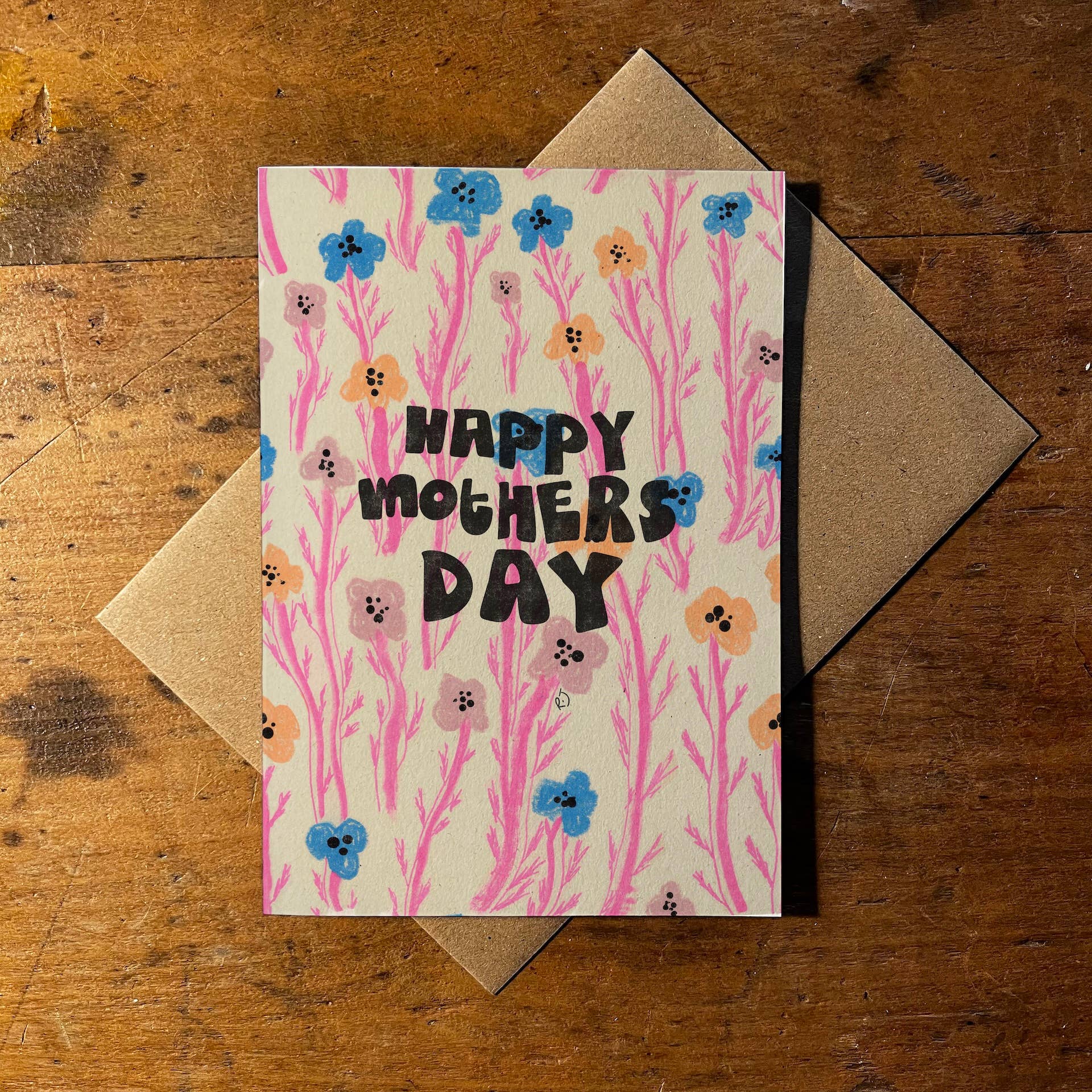 A greeting card with a floral pattern and the words 'HAPPY MOTHERS DAY' in black lettering, placed on top of a brown kraft envelope on a wooden surface.