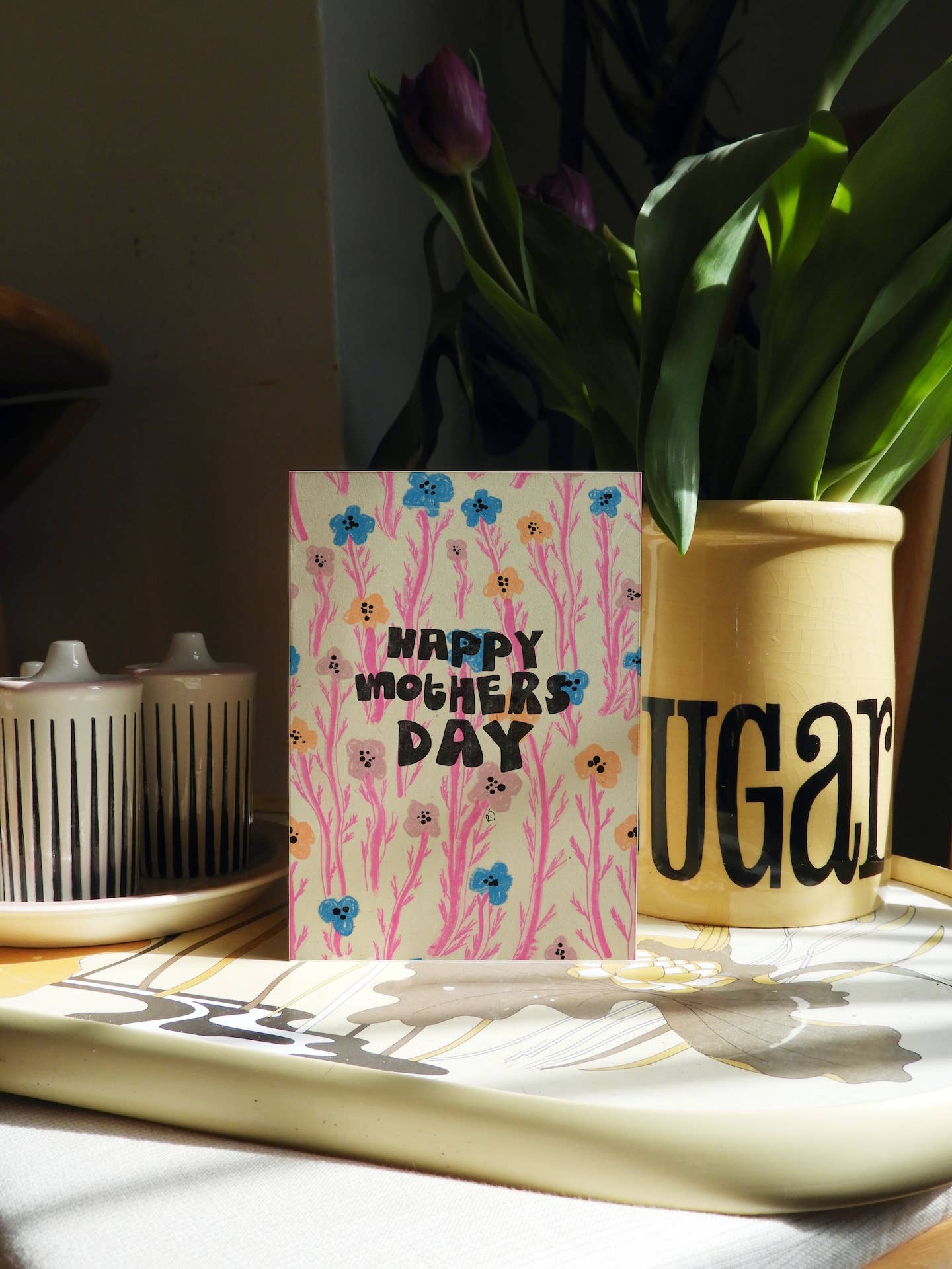 A Mother's Day greeting card with a pink floral background and the text 'HAPPY MOTHER'S DAY' in black, on a tray next to a yellow ceramic pot with tulips.