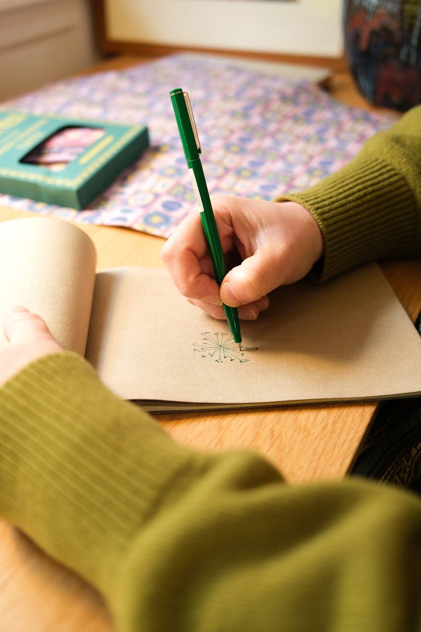 Person holding a green pen and drawing in a notebook on a wooden table.