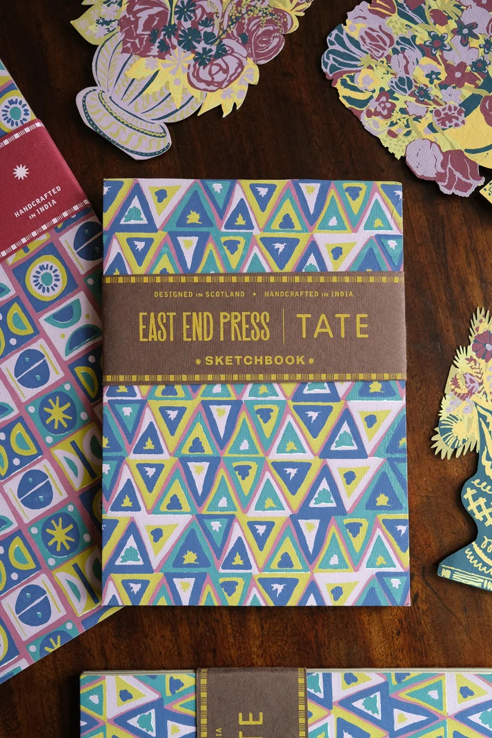 A sketchbook from East End Press x Tate, featuring a colorful geometric triangle pattern in blue, green, yellow, and purple, with a brown band across the middle. The sketchbook is displayed on a dark wooden table surrounded by other patterned stationery items.