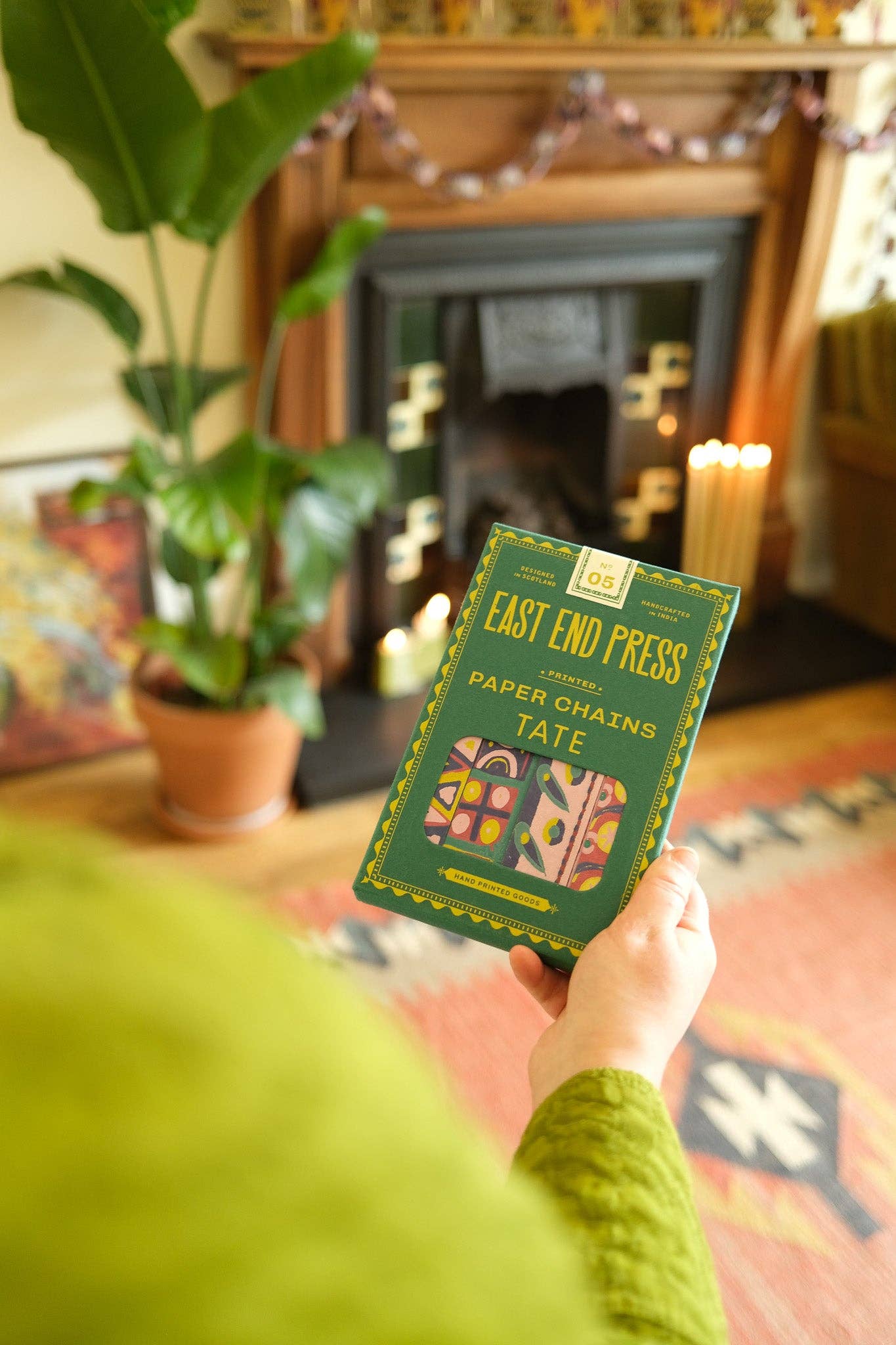 Person holding a book titled 'East End Press' in a cozy room with a fireplace and plants.