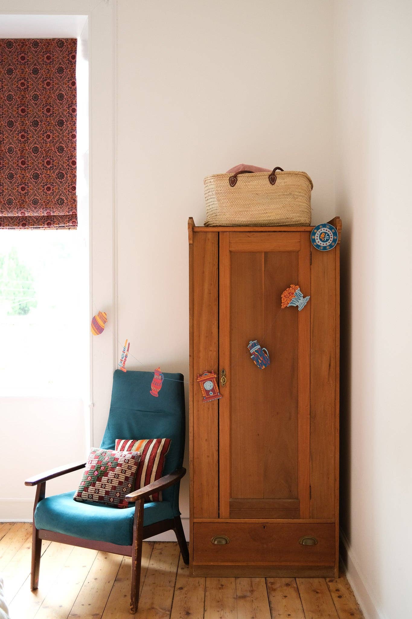 Wooden wardrobe with a basket on top next to a blue armchair with a patterned cushion, with a paper garland strung across.