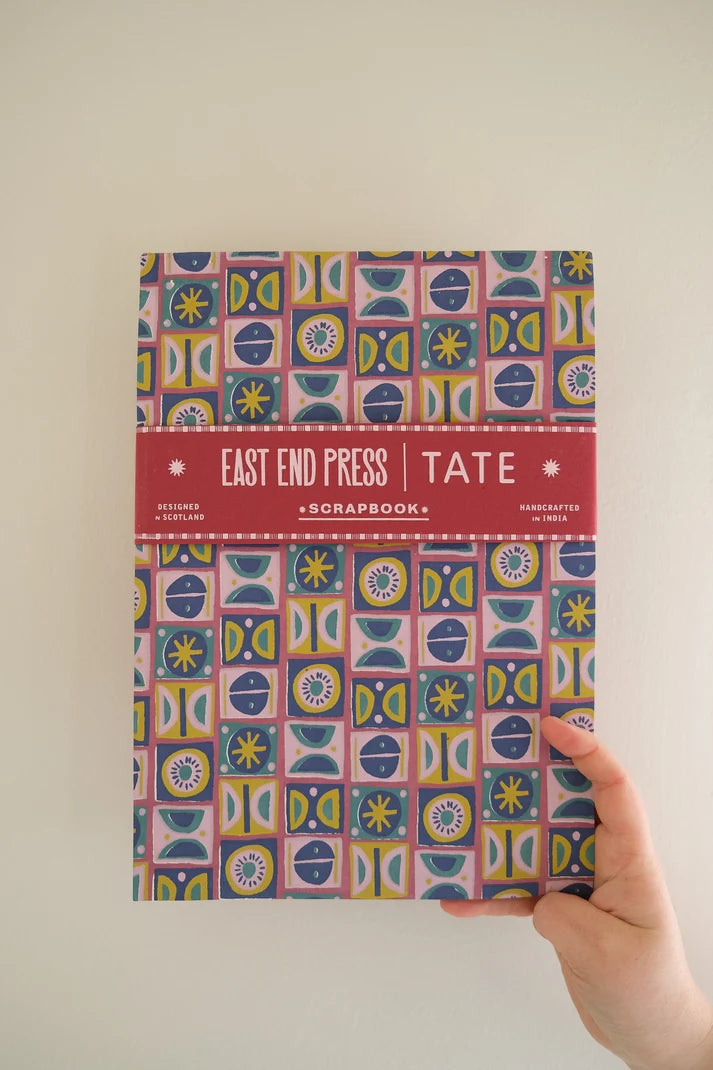 A hand holding a copy of the East End Press Tate Scrapbook against a plain beige wall.