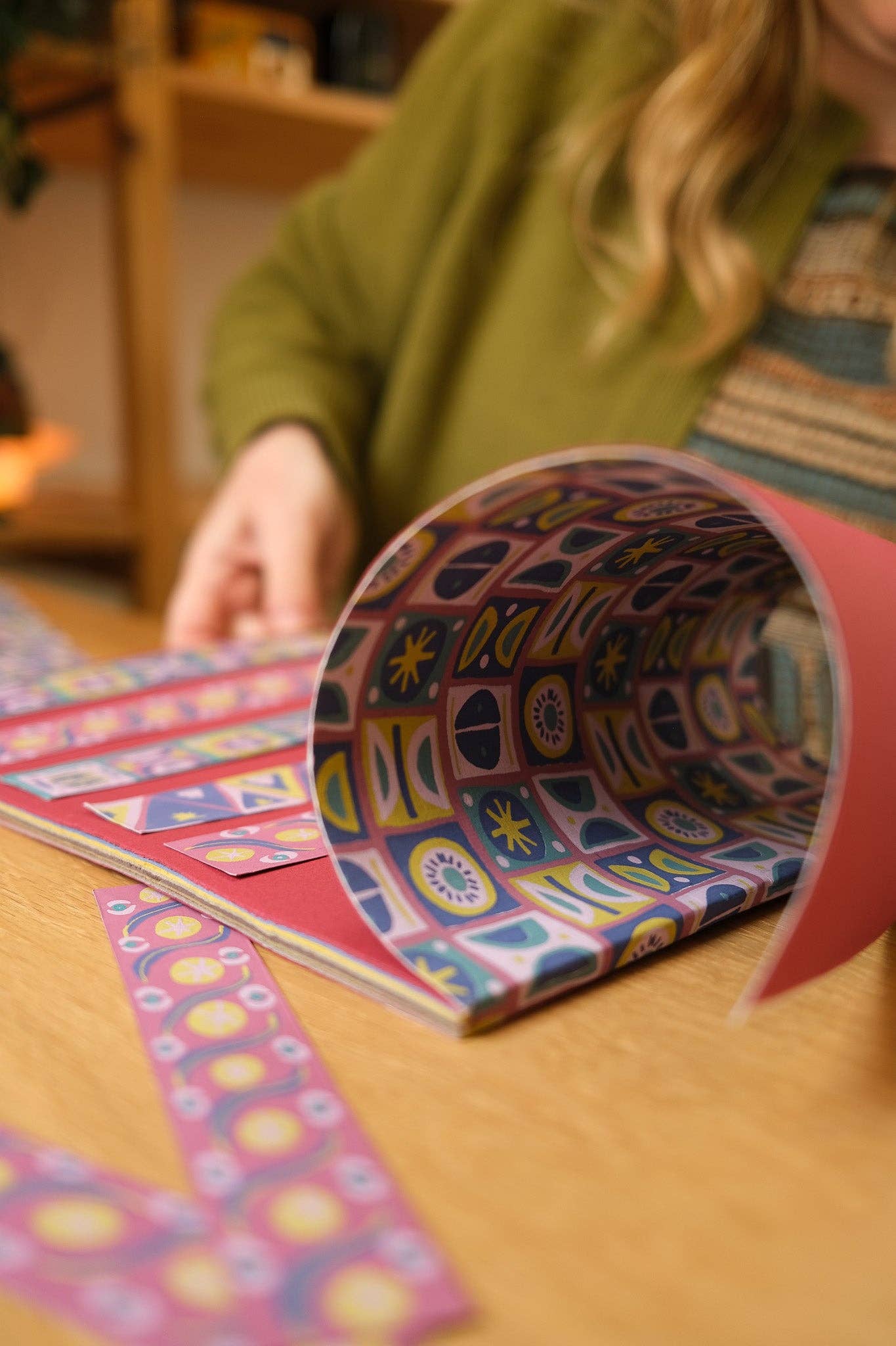 A close-up of a person rolling out a sheet of patterned pink paper on a wooden table. The paper features a colorful geometric and celestial design with stars, moons, and geometric shapes in blue, yellow, and white.