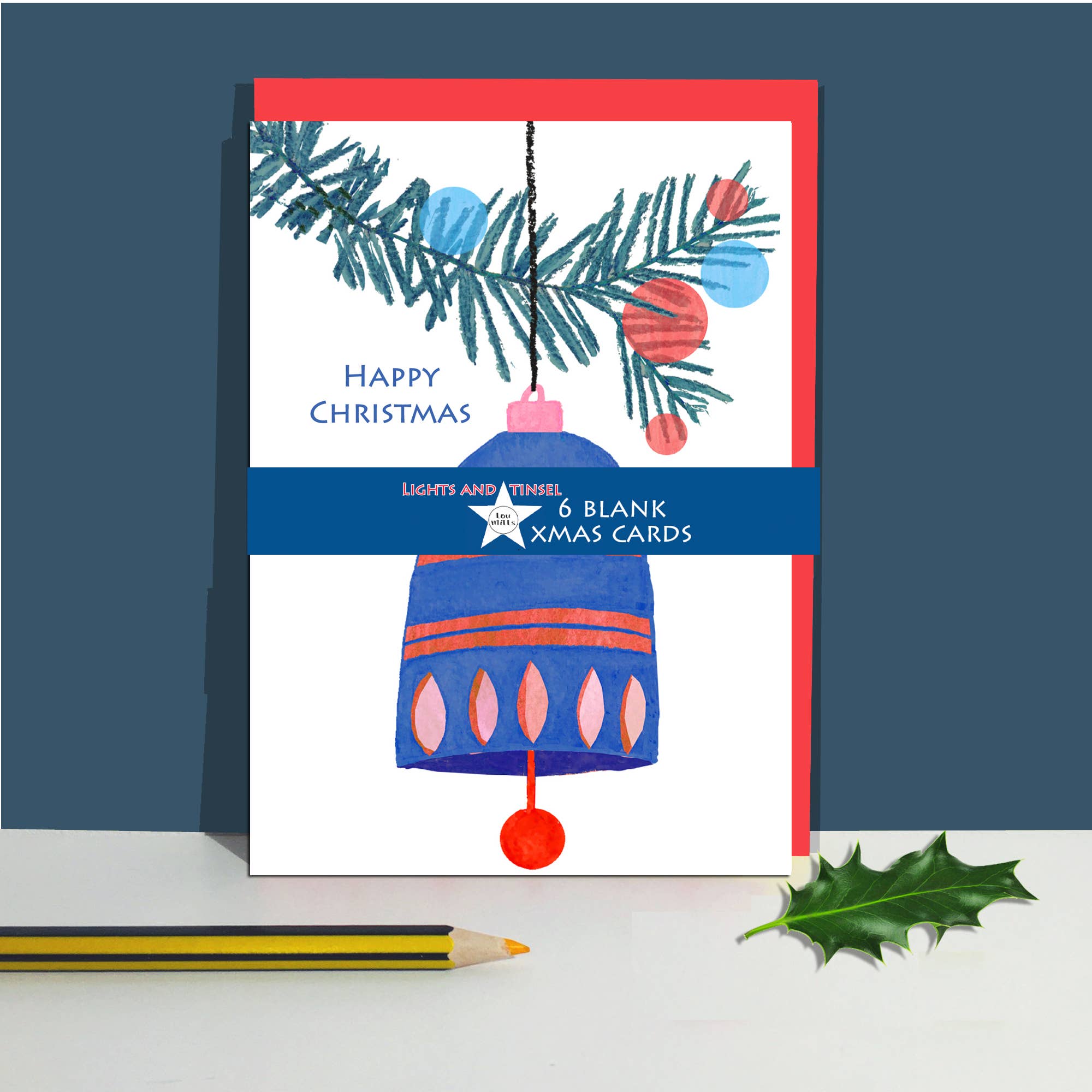 A Christmas greeting card standing upright on a white surface, with a red envelope, a blue bell illustration, pine branches, and the text 'Happy Christmas' and '6 blank Xmas cards'. A green holly leaf and part of a yellow pencil are placed beside it against a dark blue background.