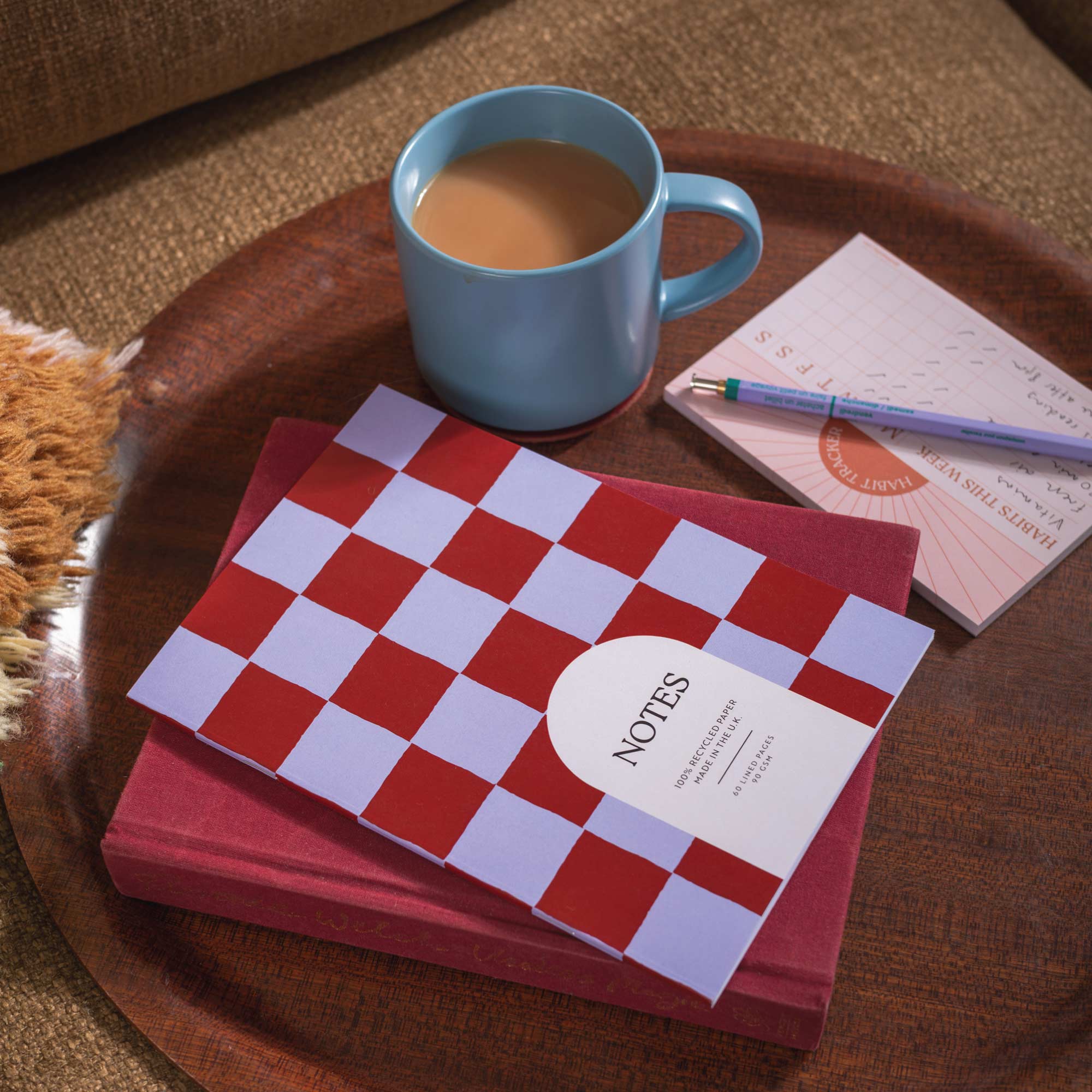 Red checkered notebook with 'NOTES' label on a wooden tray with a blue mug of coffee.
