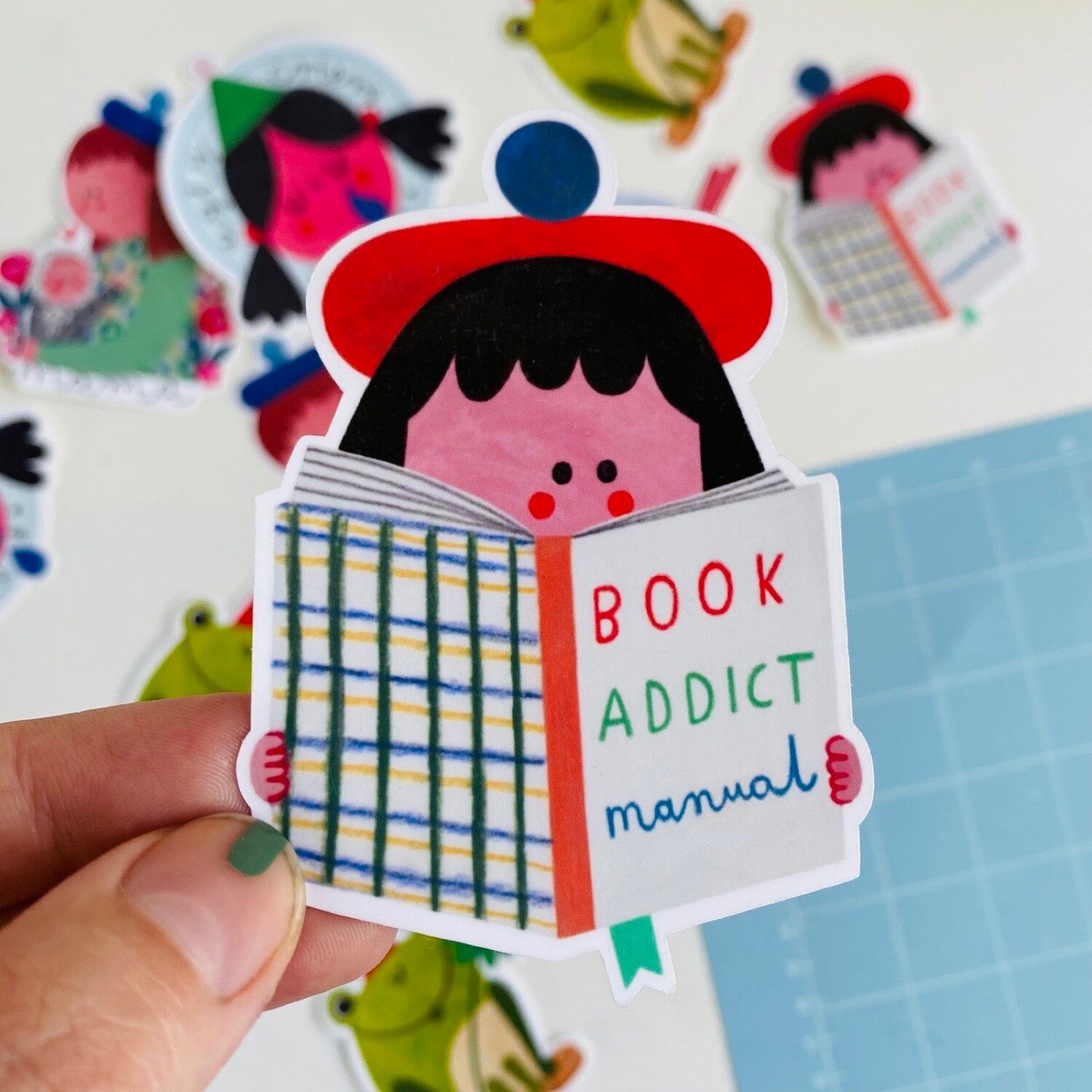 Sticker of a person reading a book titled 'Book Addict Manual' held by a hand on a blue grid background.