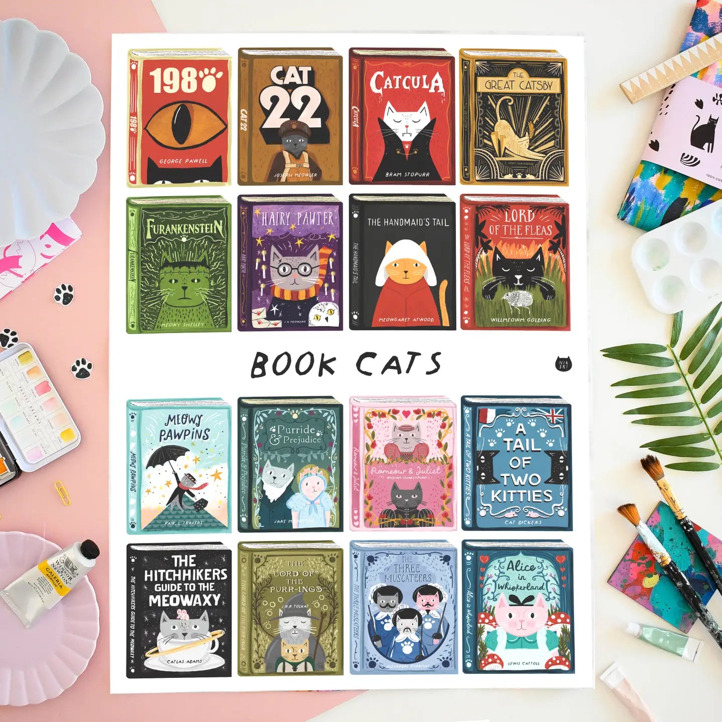 Tea towel featuring a collection of books with cat-themed covers on a white and pink background with paints around it.