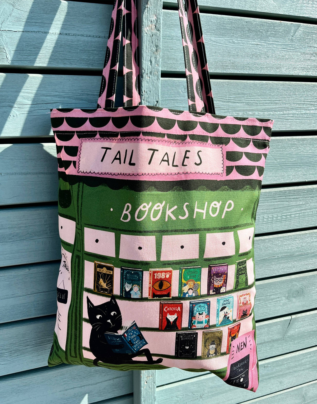 A pink and green tote bag featuring a bookshop design with the text 'TAIL TALES BOOKSHOP', illustrated book covers, a black cat reading, and patterned handles, hanging against a light blue wooden wall.