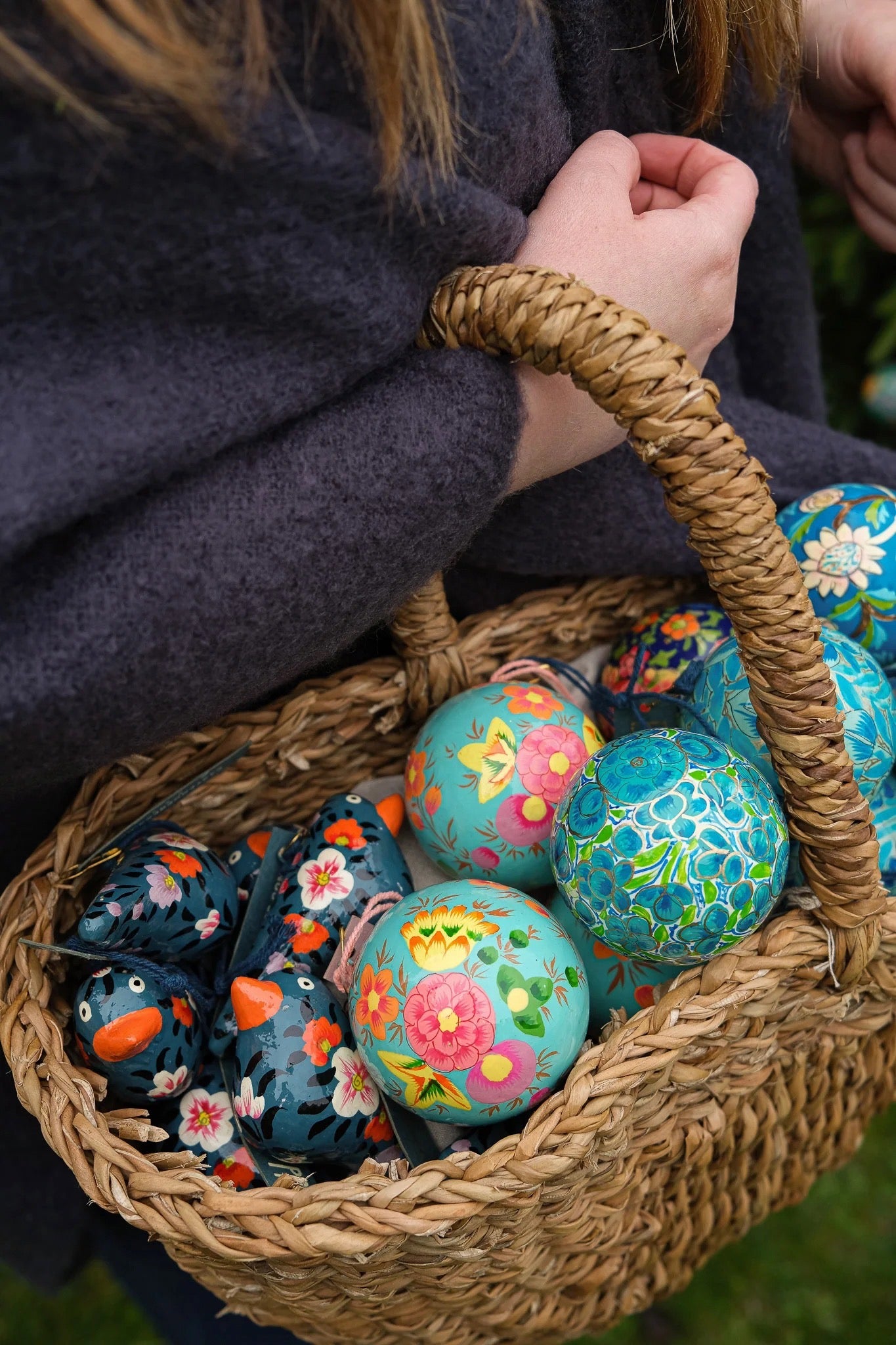 Basket filled with colorful decorated baubles held by a person wearing a dark coat.
