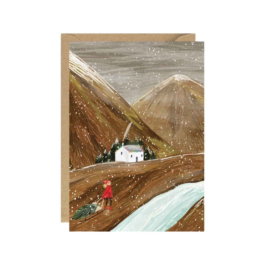 Christmas card featuring a white bothy at the bottom of the mountains and a lady in a red coat dragging a Christmas tree towards the bothy.