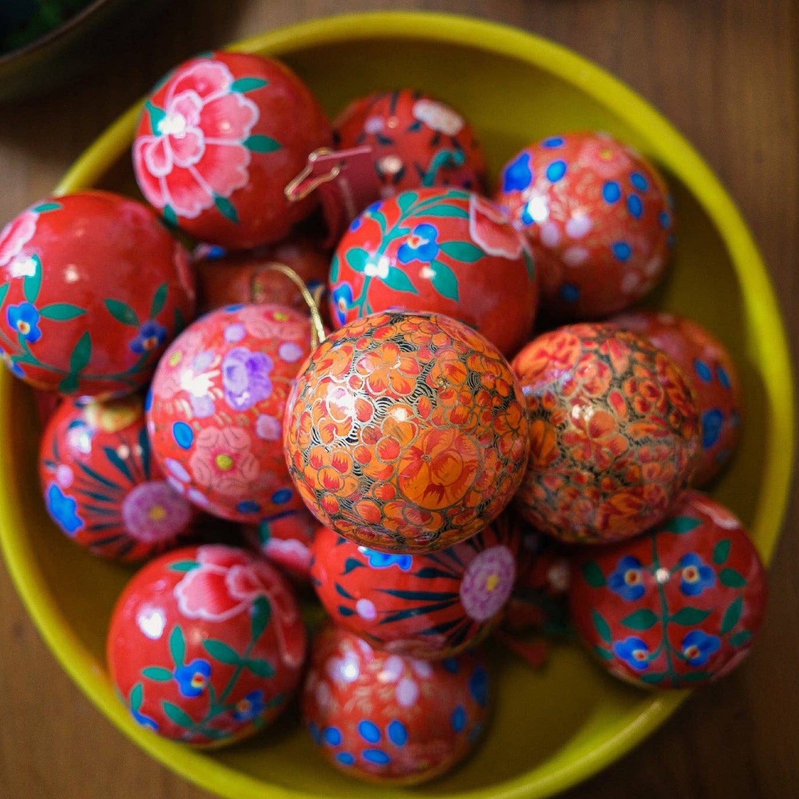 A yellow bowl of round papoer mache Christmas baubles in red floral patterns.