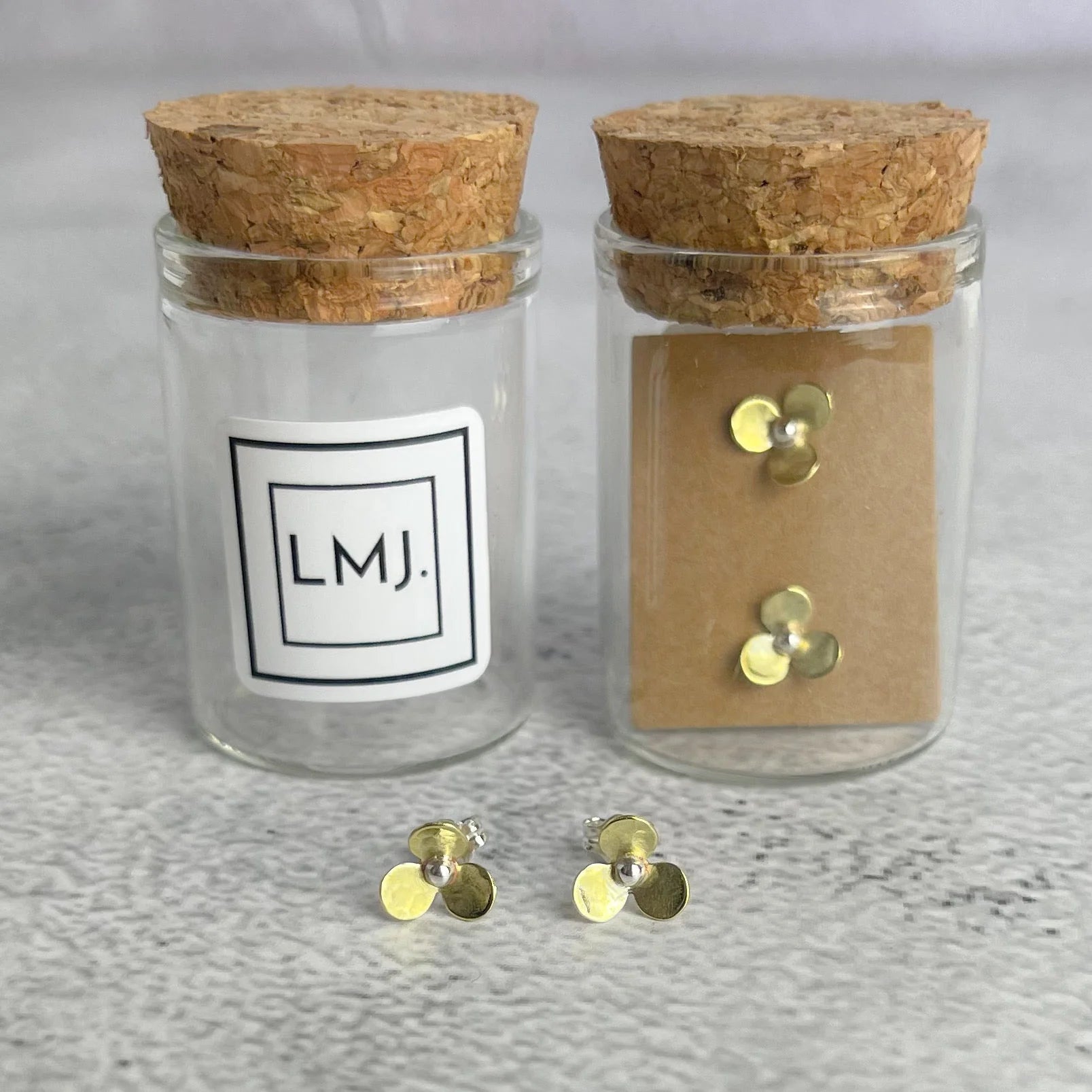 Two glass jars with cork lids, one labeled 'LMJ' and containing gold earrings, on a gray surface.