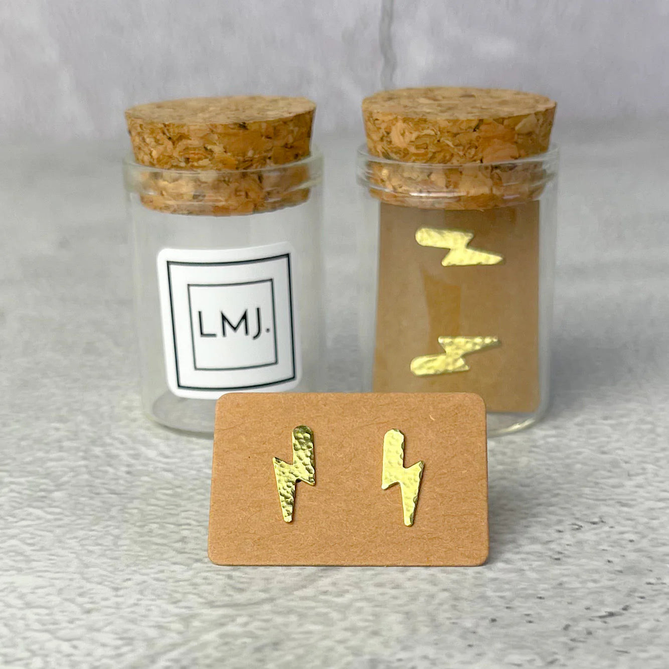 Gold lightning bolt earrings on a card with two glass jars labeled 'LMJ' in the background.